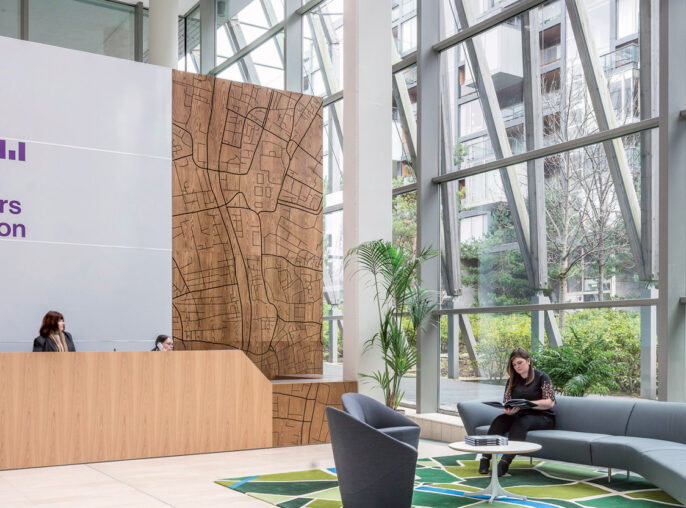 Spacious atrium lobby featuring high ceilings with geometric skylights, a large scale wood-paneled reception desk, sleek gray sofas on patterned area rugs, and airy greenery, embodying modern corporate design.