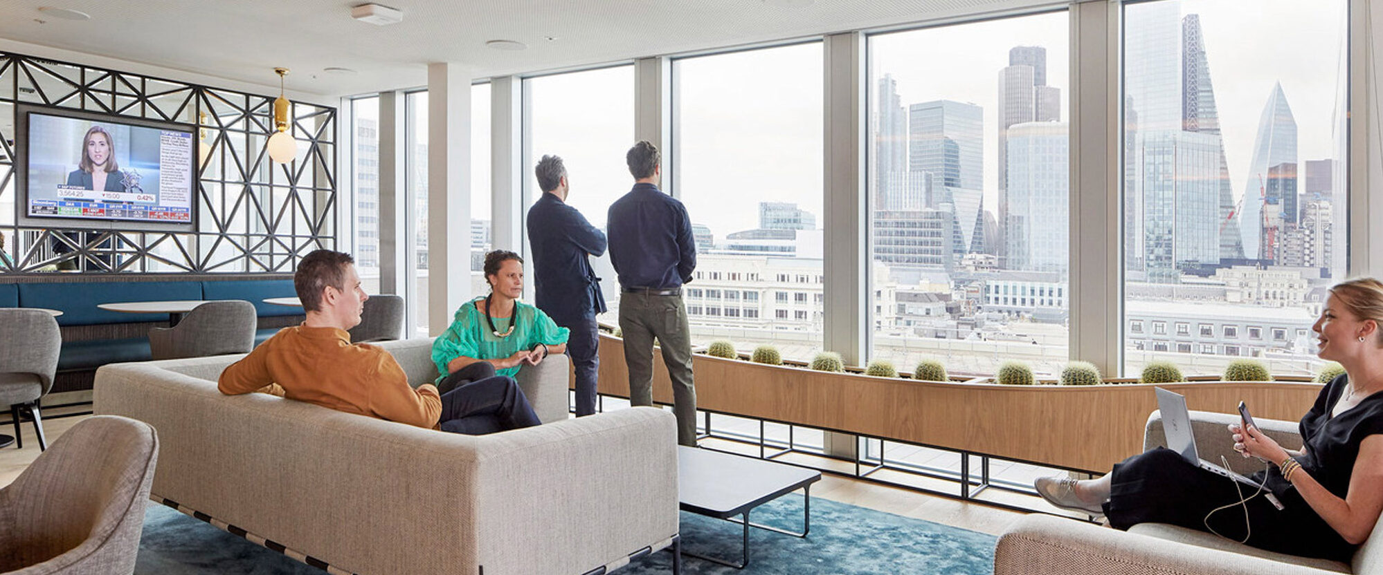 Open-plan office space with modern furniture including sofas and chairs with geometric patterns, set against large windows showcasing urban skyline views. An area rug with shades of blue anchors the seating arrangement, while individuals engage in work and conversation, under ample natural lighting.
