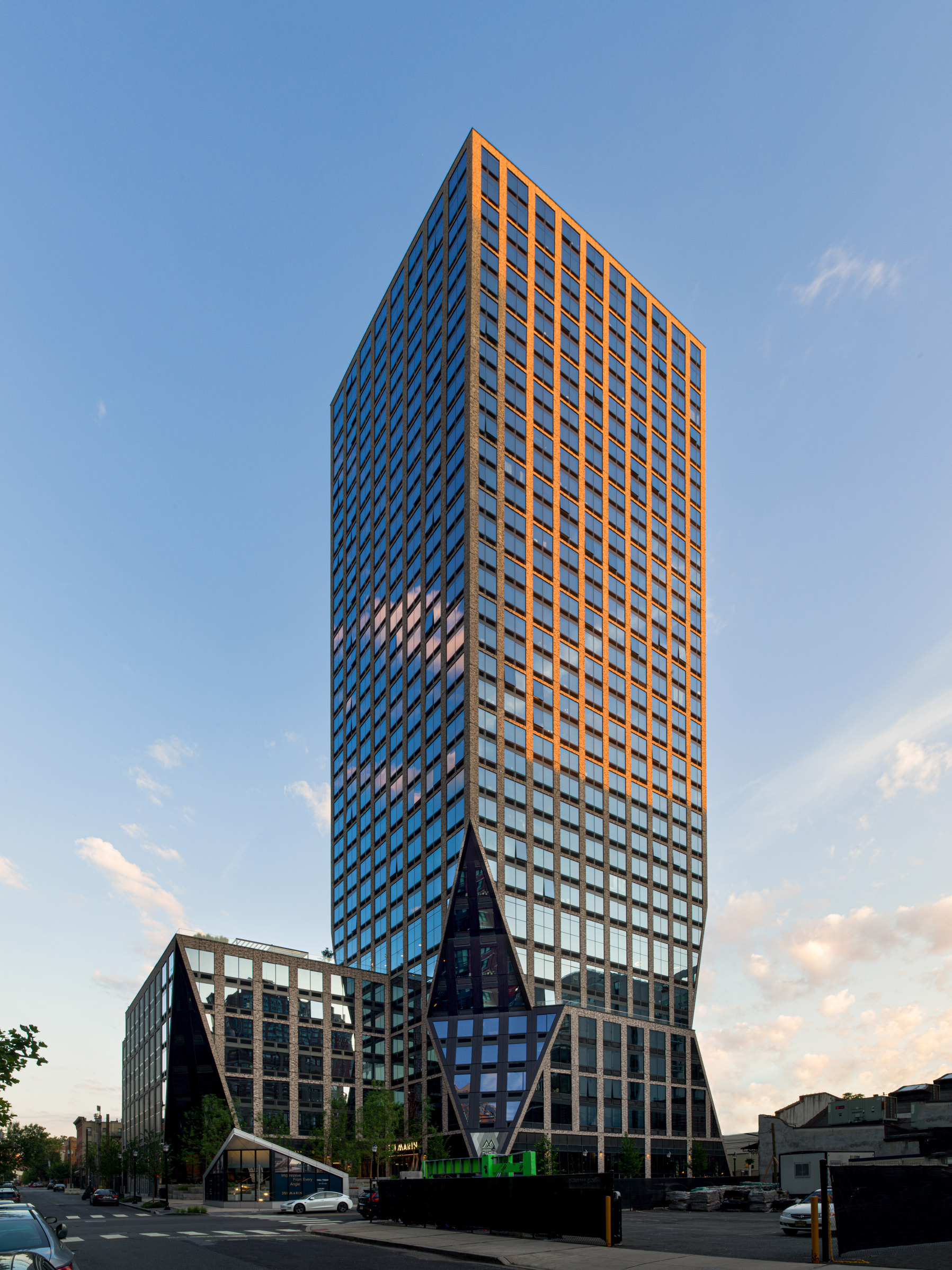 Modern skyscraper at dusk with reflective glass facade and prominent geometric lines, featuring a juxtaposed lower structure with a triangulated design element, showcasing contemporary architectural innovation in an urban setting.