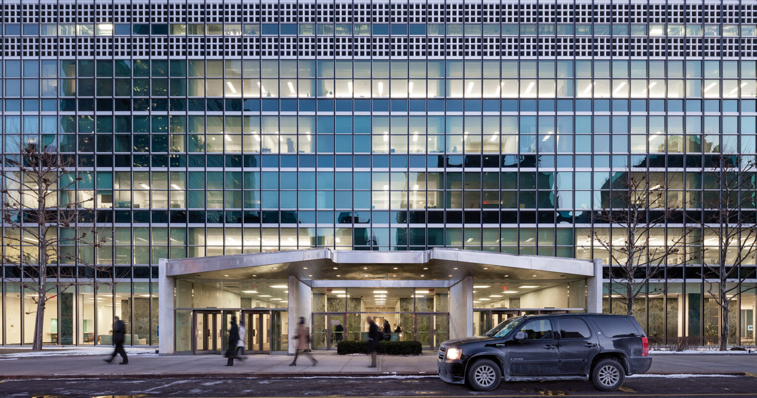 Modern office building facade with pedestrians and a vehicle on the street at twilight.