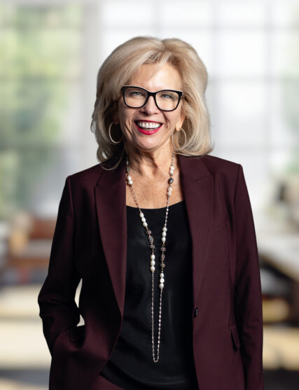 The image features a person wearing a dark blazer and a necklace, standing. The individual is set against a blurred background of an indoor setting.