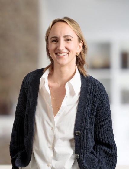 The image shows a woman with shoulder-length blonde hair and a warm smile, wearing a white blouse and a navy blue cardigan. The soft-focus background suggests a bright and spacious interior, possibly an office or gallery space.