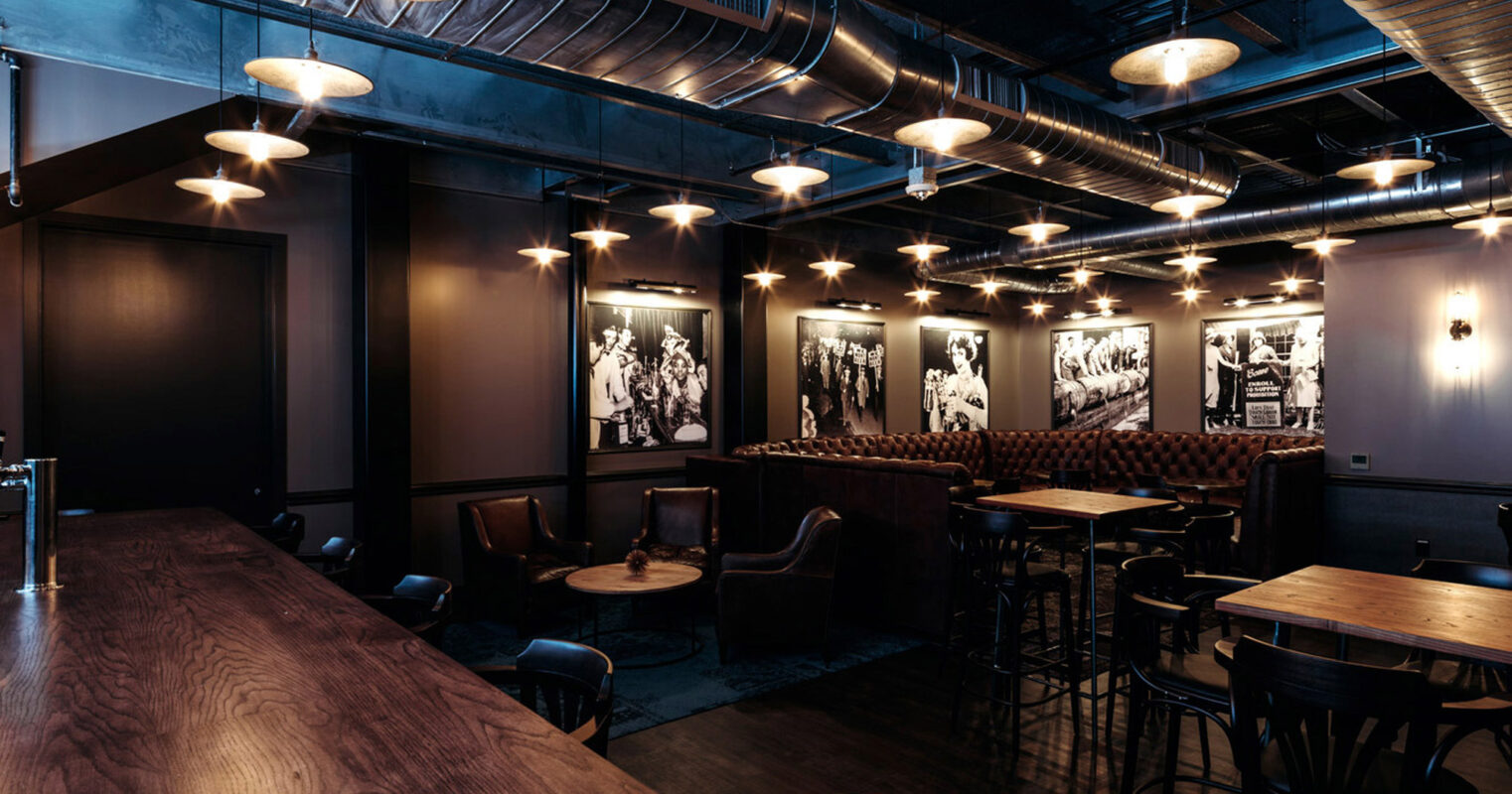 Sophisticated lounge area featuring dark wood tones and leather seating. Exposed ductwork on the ceiling contributes to the industrial-chic ambiance, while round pendant lights add warmth. Monochrome photographs line the walls, complementing the elegant, moody aesthetic.