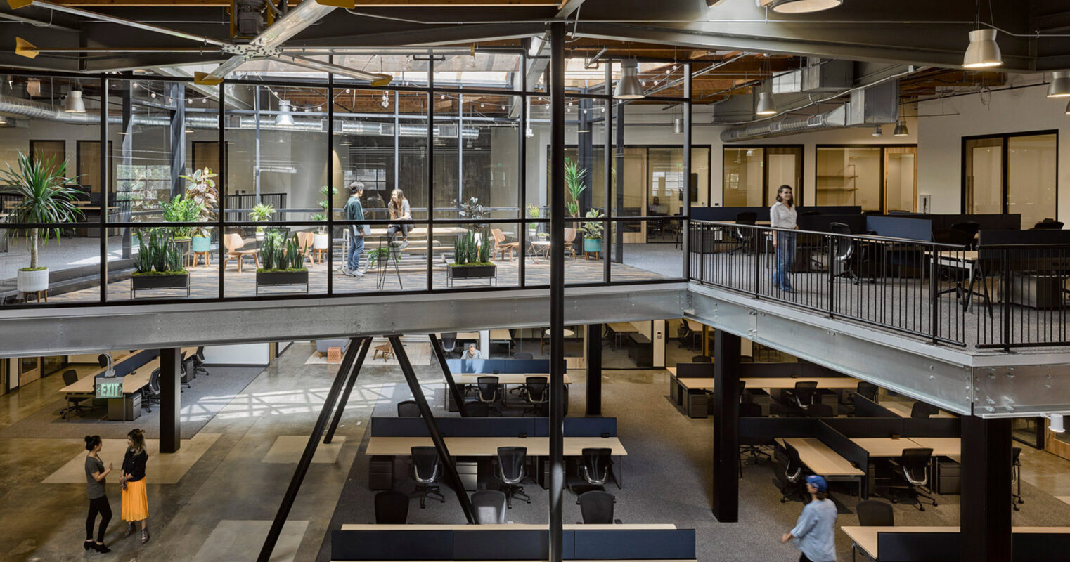 Open-plan office space featuring exposed beams and HVAC ductwork, with wood-toned desks, black ergonomic chairs, and pendant lighting. Glass-walled meeting rooms and abundant greenery add to the modern, functional design promoting collaboration and airflow.