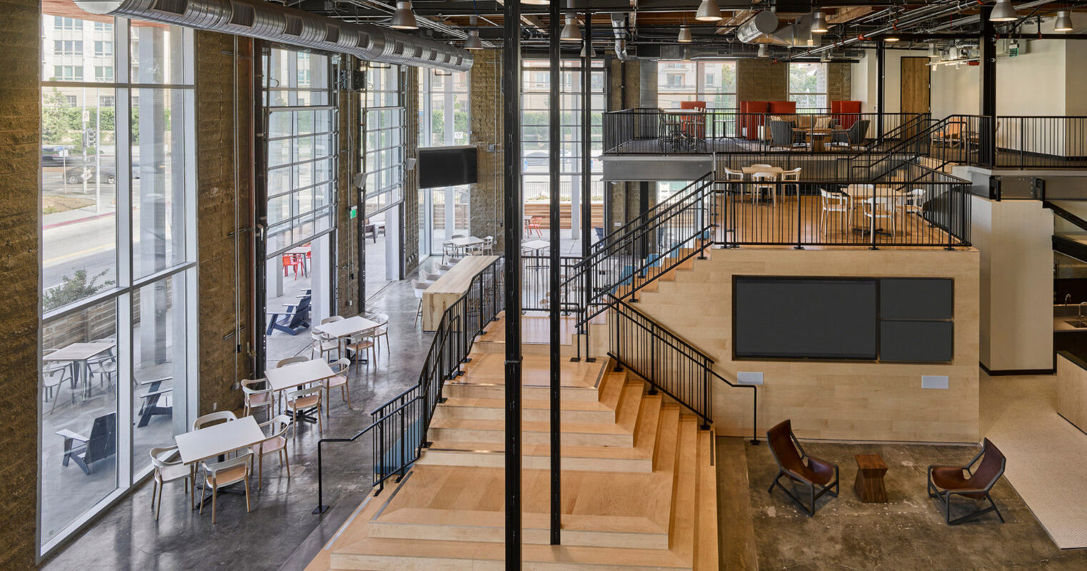 Open-plan office space featuring exposed ceiling beams and ductwork, complemented by industrial-style pendant lighting. A central wooden staircase with metal railings leads to a mezzanine level with additional seating areas. Large windows allow ample natural light, highlighting the modern, minimalist furniture arrangement.