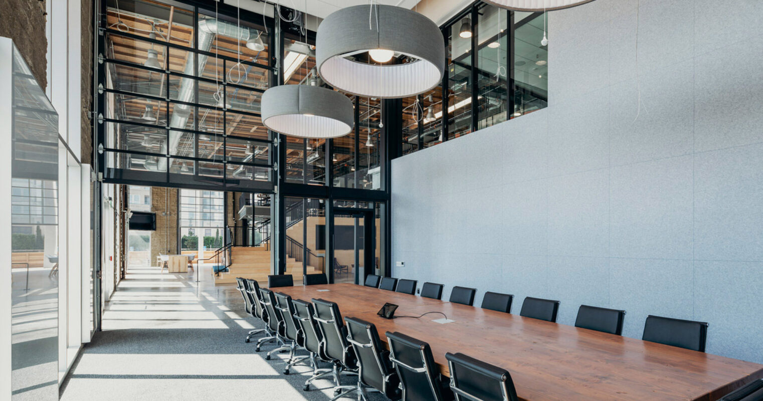 Modern conference room featuring a long, dark wood table with black chairs, complemented by large, circular pendant lights. Expansive windows allow natural light to illuminate the minimalist decor and textured walls, enhancing the room's sleek, contemporary aesthetic.