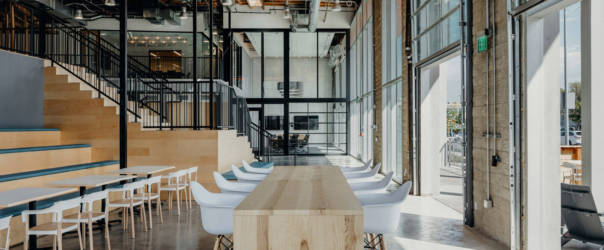 Spacious modern industrial office with high ceilings, exposed ductwork, and a mix of natural wood elements. Minimalist furniture includes sleek white tables and streamlined wood benches. Large windows provide ample natural light, complementing the warm wooden flooring and staircase.