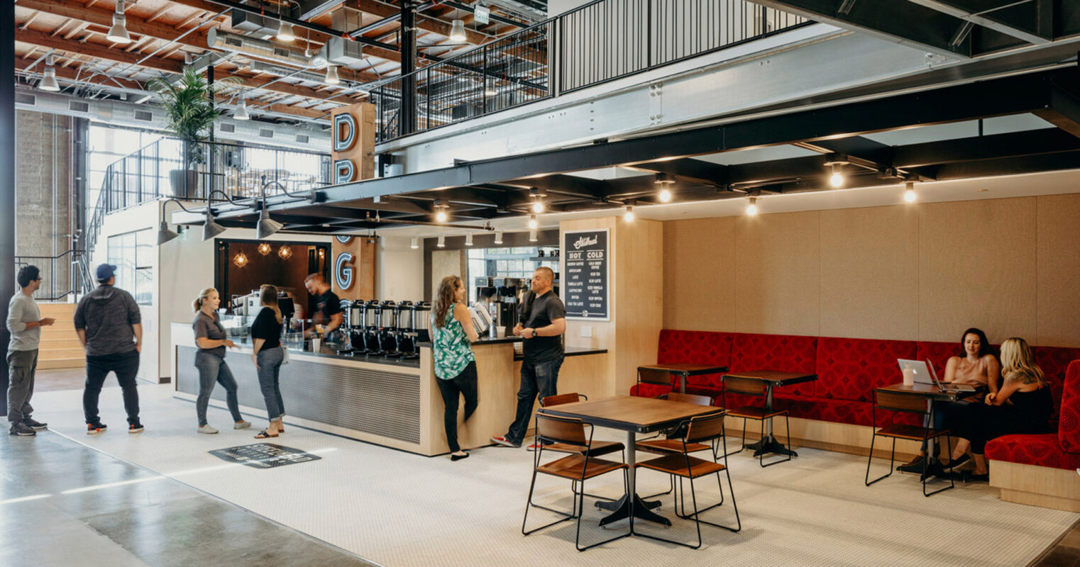Modern industrial-style coffee shop featuring exposed ceiling pipes, mixed-use concrete, and wood flooring. Suspended linear lighting accentuates the bar area, while booth seating with red upholstered benches offers intimate gathering spaces. A mezzanine level overlooks the bustling space below.