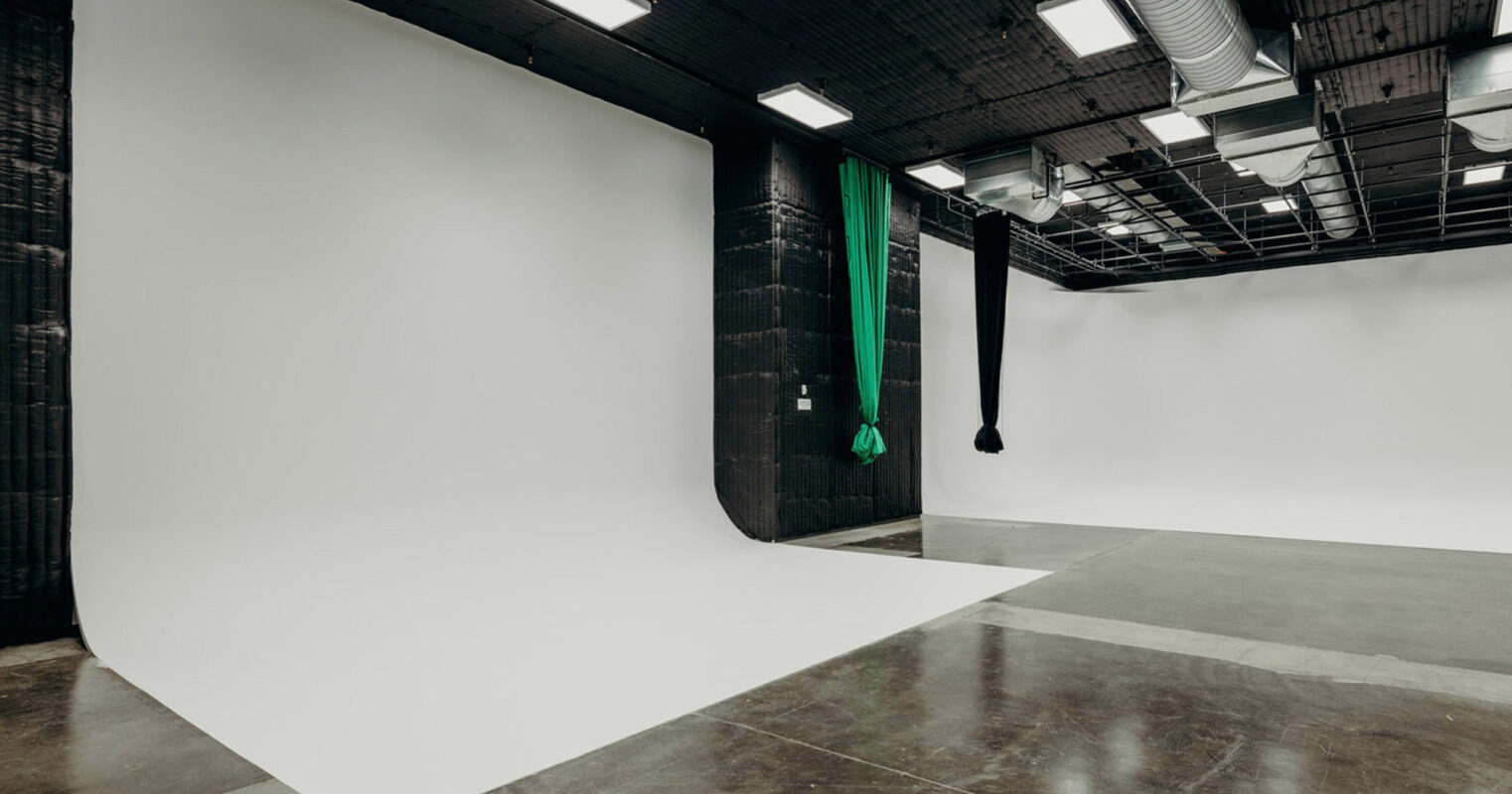 Spacious modern photo studio with a large cyclorama wall. The studio features high ceilings with exposed ductwork, concrete floors, and a harmonious blend of black and white walls for a minimalistic look. A green velvet curtain adds a touch of color and texture to the space.