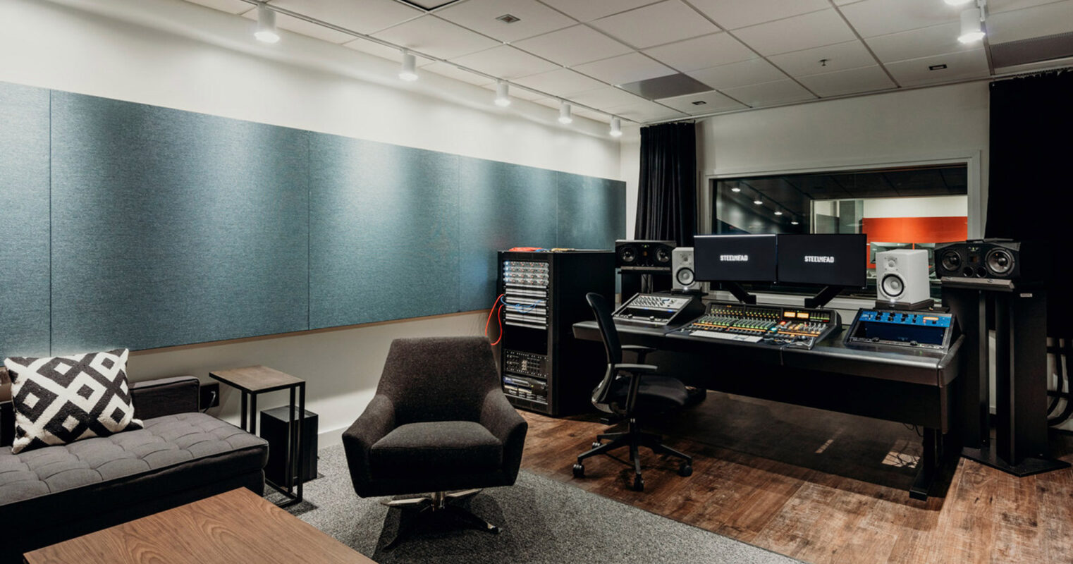 Modern recording studio with acoustic panels on walls, mixing console center, professional monitors on stands, and an ergonomic chair. Wood flooring transitions to carpet, enhancing the room's acoustics, with a cozy sofa and patterned cushions to the side for guests.