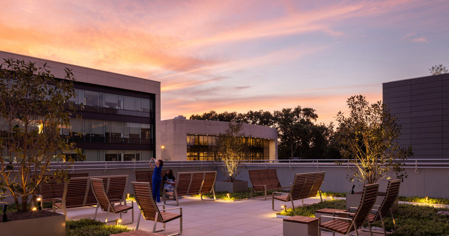 Rooftop terrace at dusk with integrated wooden seating and planters, under-floor lighting, against a backdrop of modern architecture and a vibrant sunset sky.