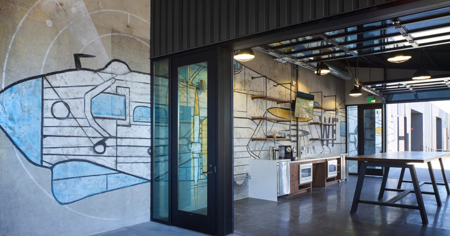 Contemporary industrial-style office space featuring exposed ductwork, polished concrete floors, and a large graffiti mural adding an artistic accent to the textured gray walls. Glass doors, sleek furnishings, and pendant lighting complete the modern aesthetic.