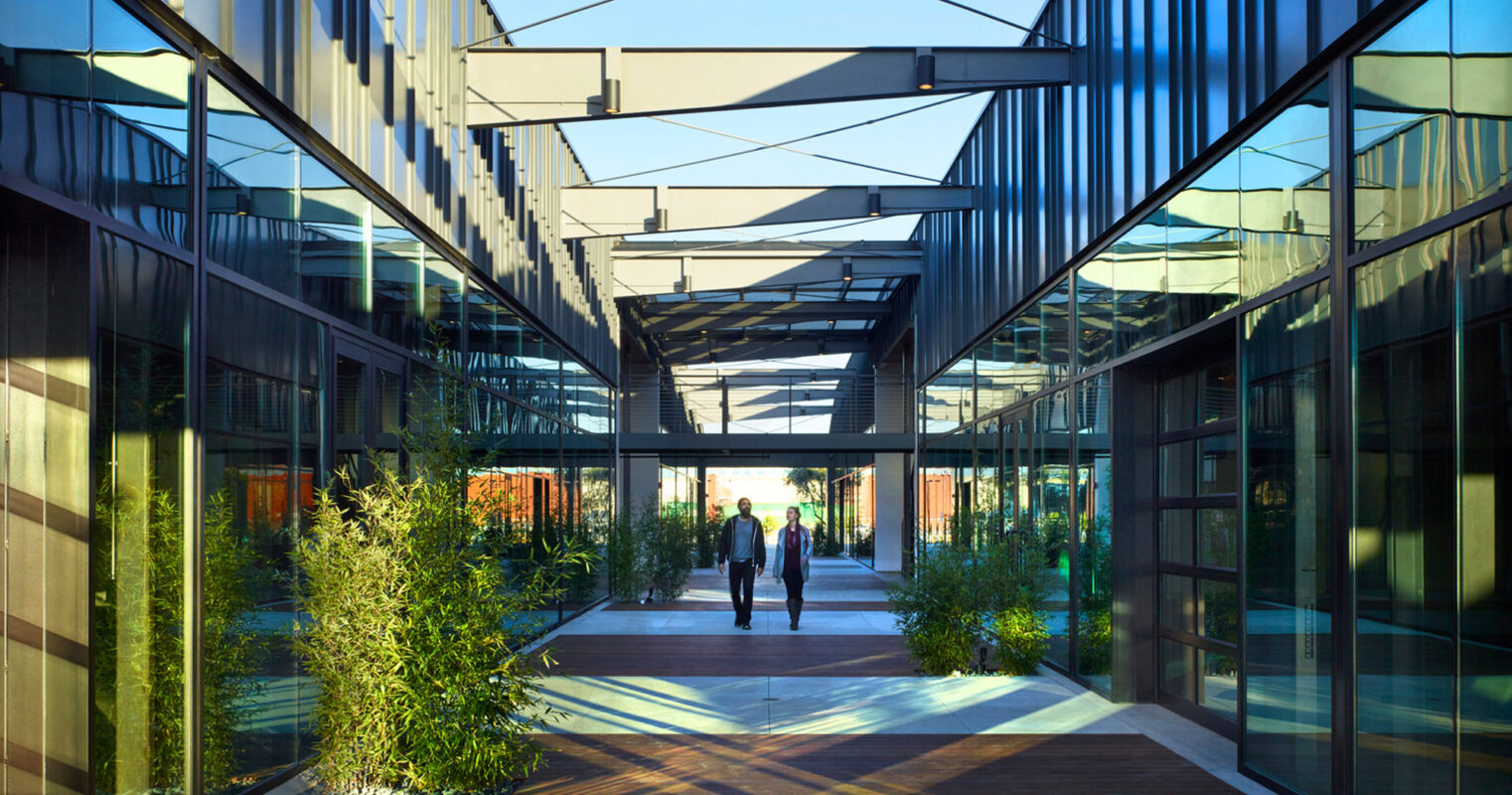 Modern corporate atrium with structured glass walls, reflecting sunlight and sky. Overhead beams and cables add an industrial feel, while green plant beds infuse a natural touch into the geometric space. Pedestrians stroll along the wooden pathway, adding a sense of scale and human interaction.