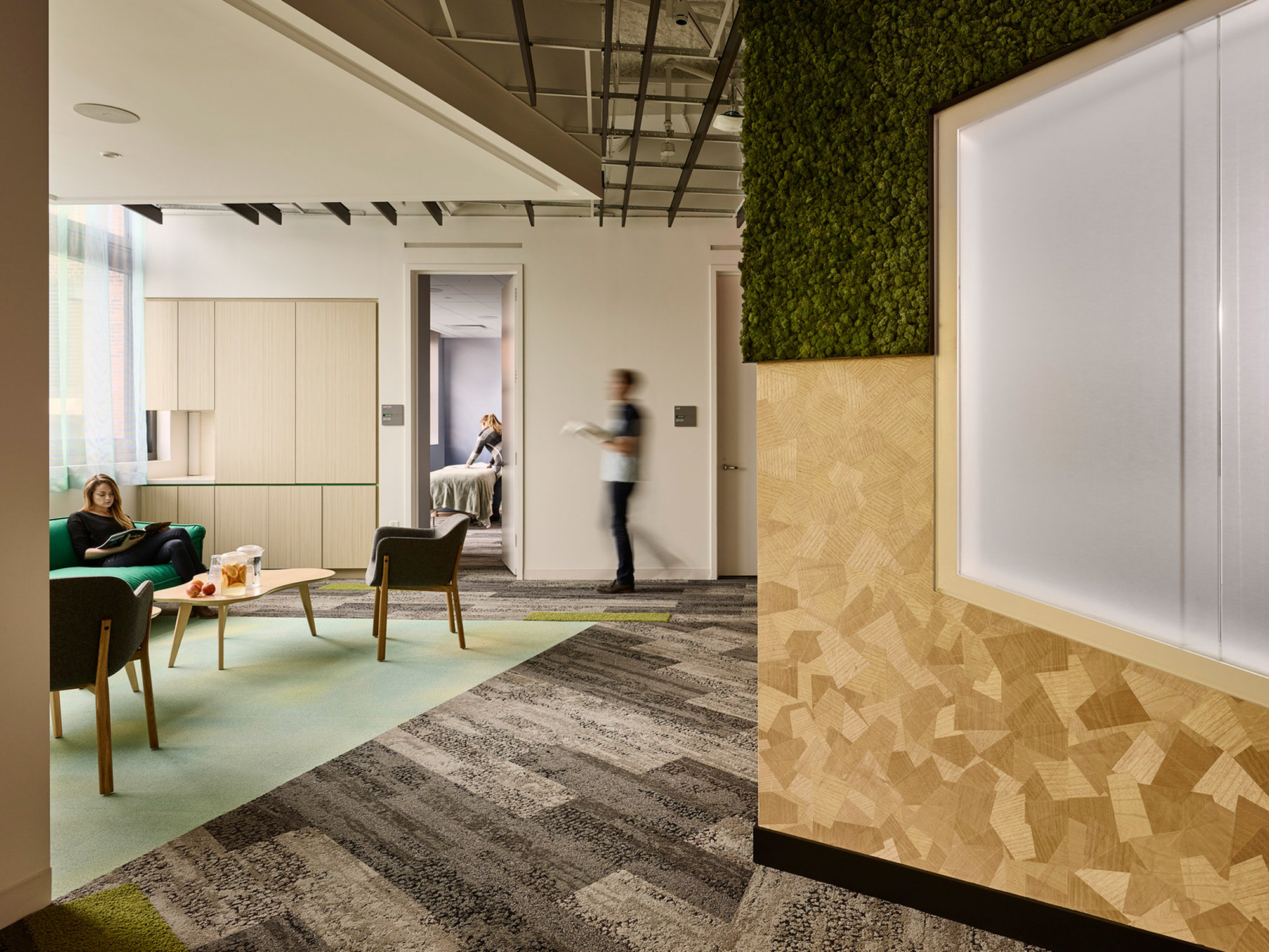 Modern office lounge with herringbone wood wall, integrated greenery, and contemporary furnishings on mixed-pattern carpeting. Natural light illuminates the relaxed workspace.