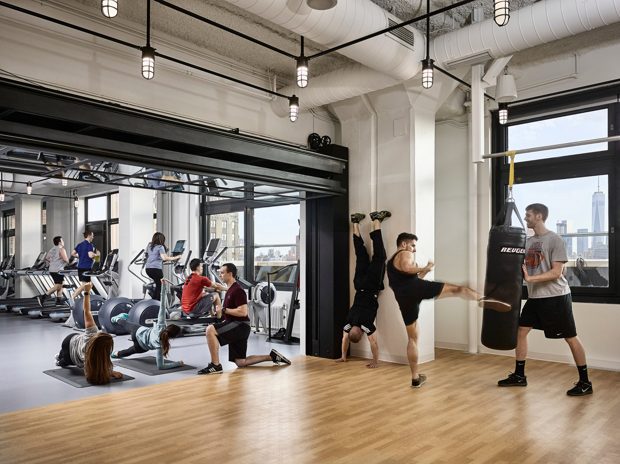 Modern fitness center showcasing expansive floor-to-ceiling windows with city views, exposed ductwork for an industrial aesthetic, and a variety of exercise equipment for cardio and strength training. Several individuals are engaged in workout activities, creating a dynamic and energetic atmosphere.