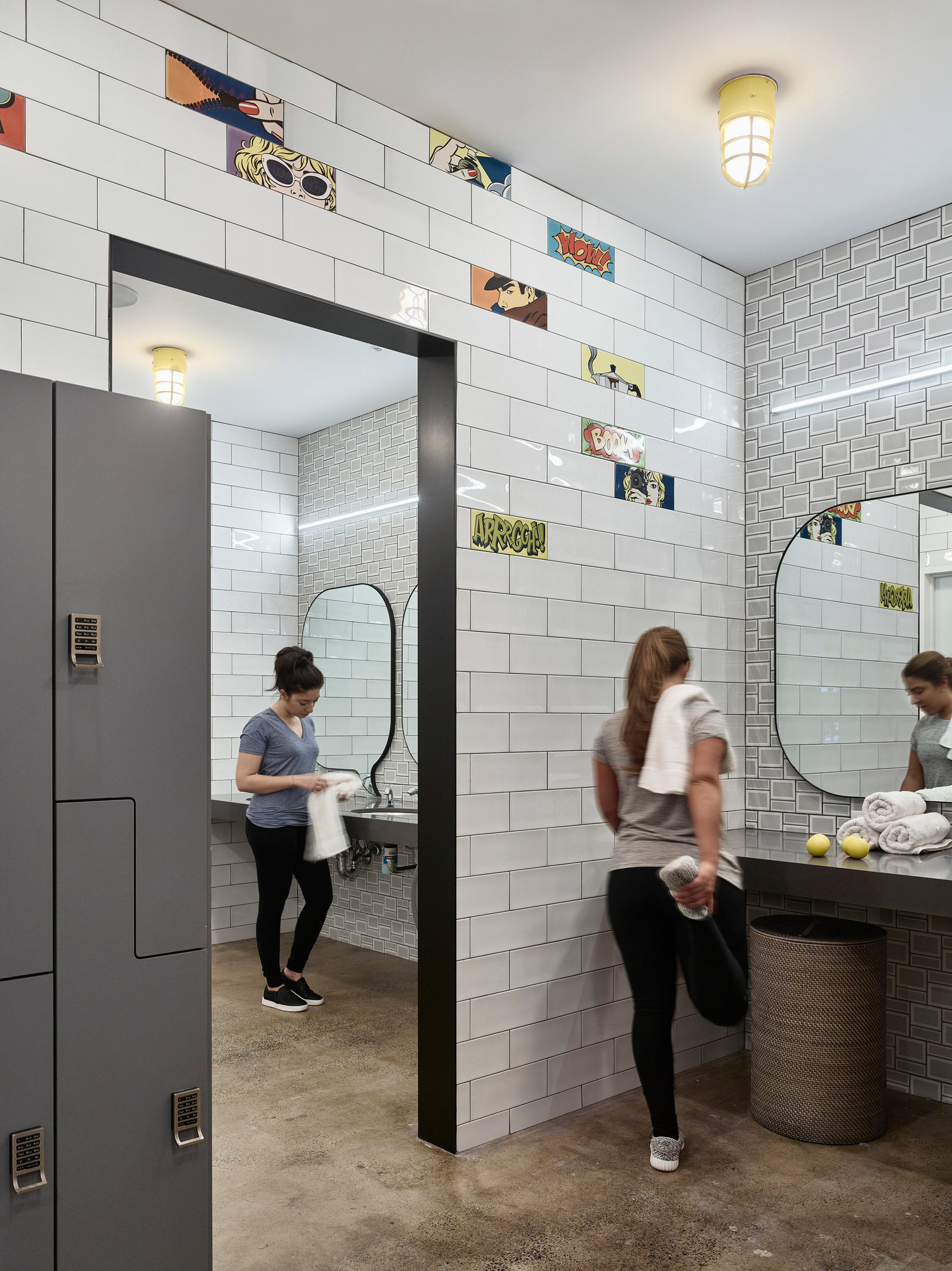 Modern restroom design with white subway tiles walls and decorative border featuring colorful illustrations. Twin circular mirrors hang over a sleek basin counter, while an industrial-style black doorframe contrasts the space. The contemporary aesthetic is complemented by warm, diffused lighting from pendant lamps.