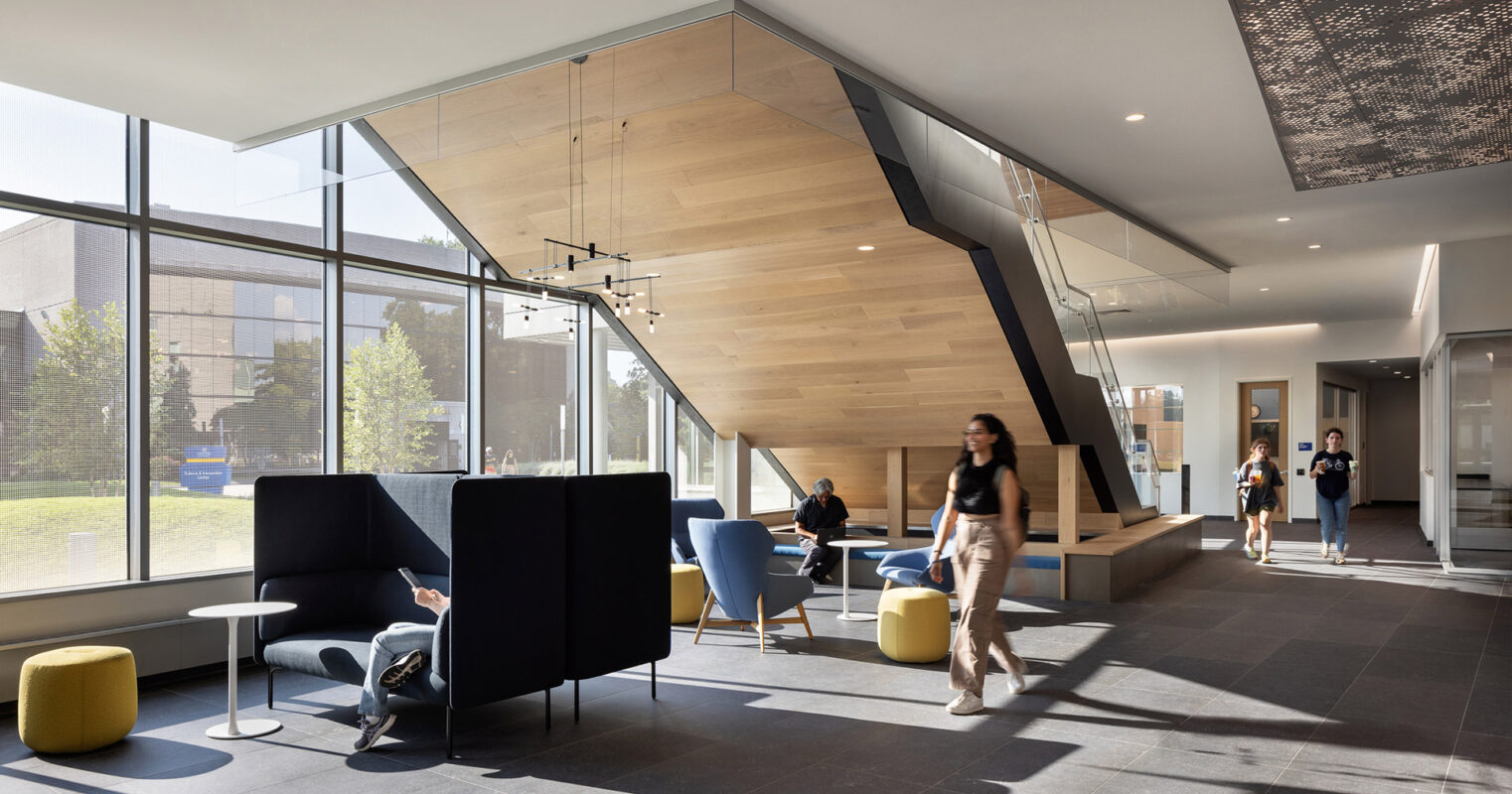 Warm natural light floods a modern lobby with a geometric wood-paneled staircase as the centerpiece. Sleek, dark furniture contrasts with the light floor, accented by vibrant yellow ottomans. Expansive glass walls offer a seamless indoor-outdoor connection.