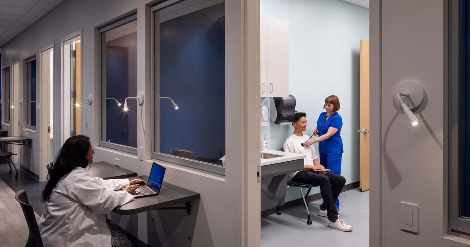 Modern healthcare office with ergonomic design featuring a row of cubicles with glass partitions, sleek white desks, and blue privacy curtains. Healthcare professionals engage with patients, reflecting a dynamic and client-centered environment.