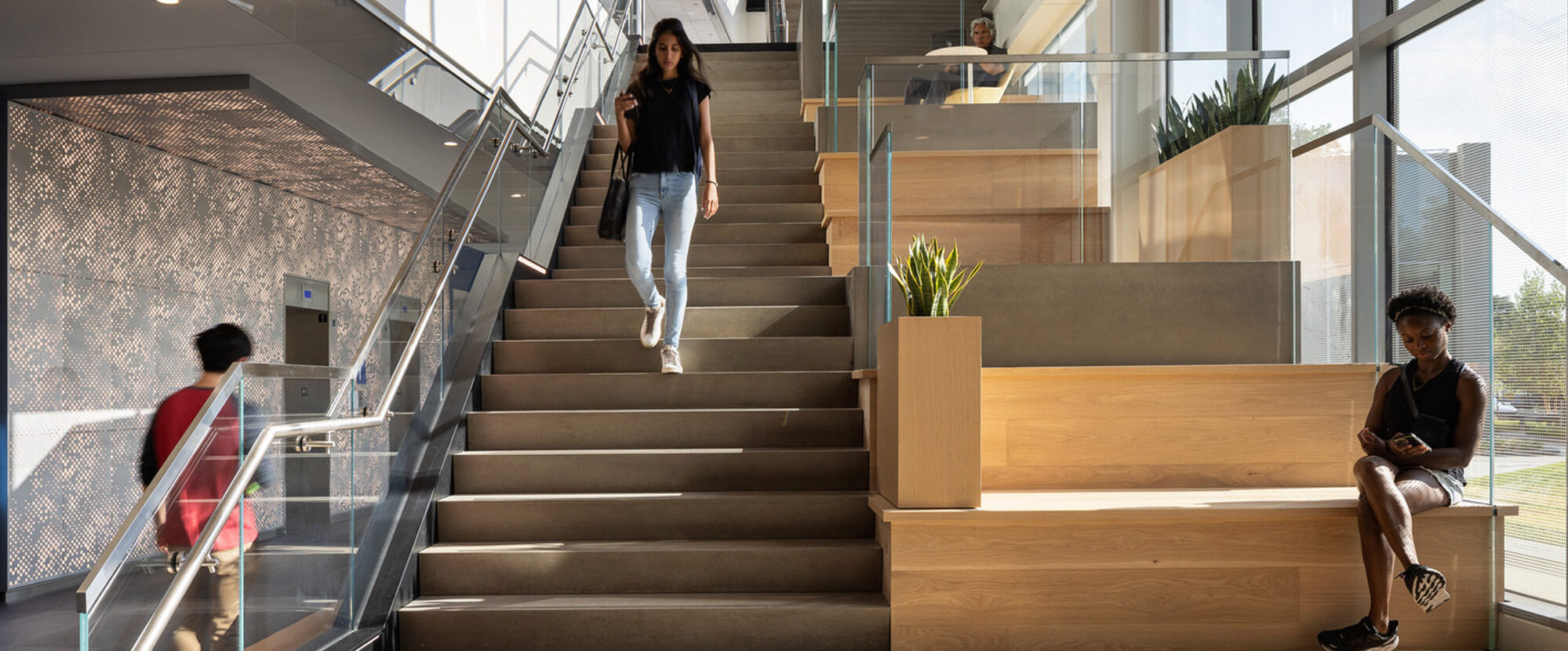 Modern staircase intersects with wooden seating steps in a light-filled atrium, as individuals engage with the multi-functional space, showcasing a blend of functionality and design aesthetics.