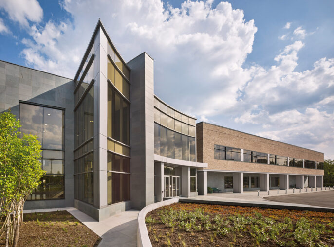 Modern architectural exterior featuring a juxtaposition of sleek, reflective glass and matte stone finishes. Angled rooflines and a mix of vertical windows create a dynamic facade, with landscaped greenery complementing the structure's geometric precision.