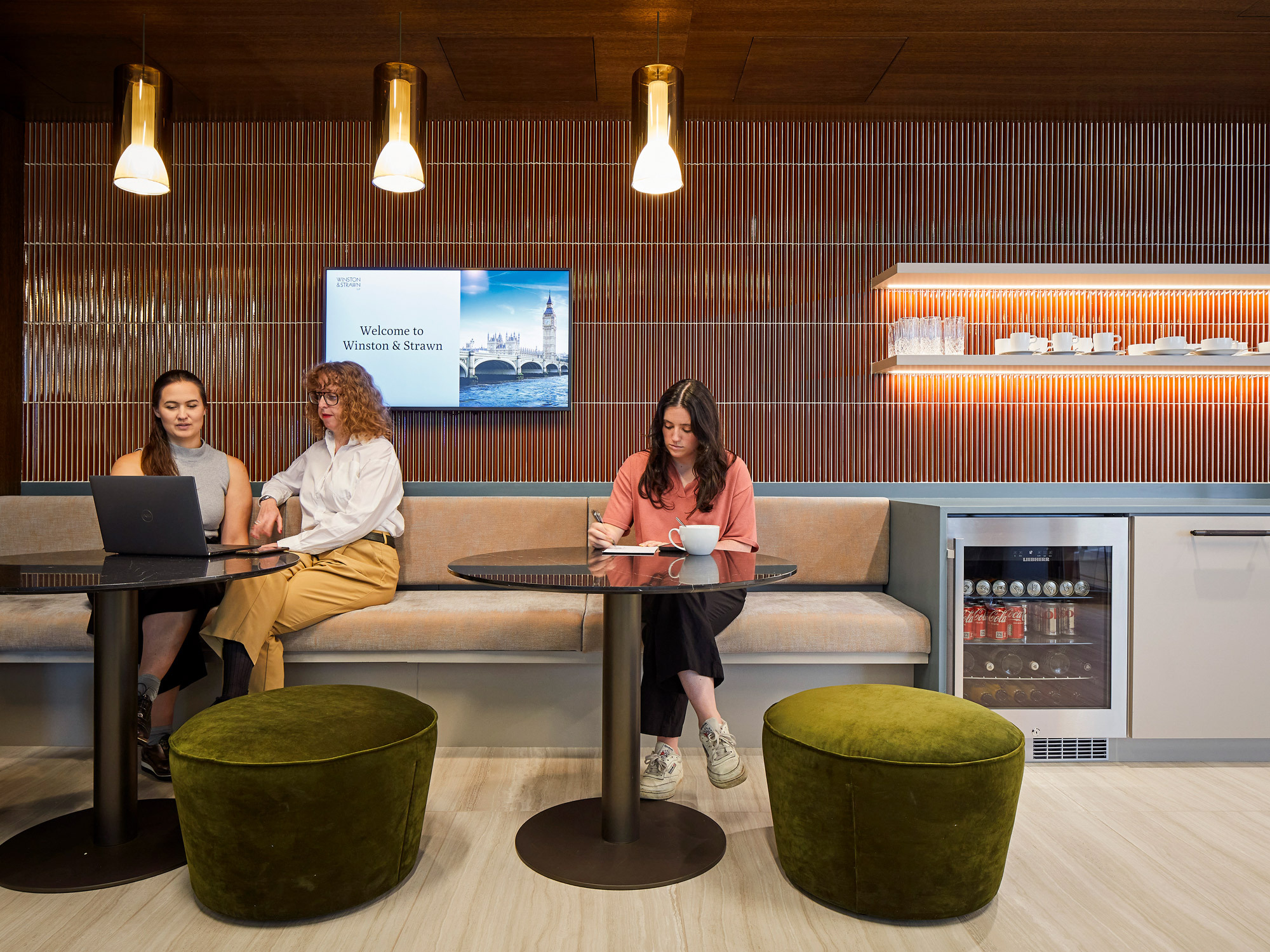 Modern office lounge with a warm wooden slat wall, complemented by sleek hanging pendant lights. A digital welcome sign adds a corporate flair, while plush green ottomans offer casual seating, beside a tidy kitchenette with a beverage fridge.
