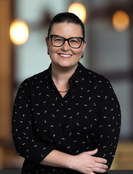 A smiling woman with shoulder-length dark hair poses for a professional portrait. She is wearing large black-framed glasses, a black blouse with a small white pattern, and small hoop earrings, conveying a friendly and approachable demeanor.