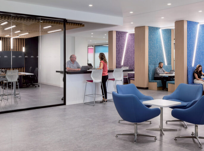 Contemporary office break room with vibrant blue egg chairs, sleek white high-top tables, and people in casual conversation. Textured columns with purple accents complement the neutral-toned flooring and wooden slatted ceiling design.