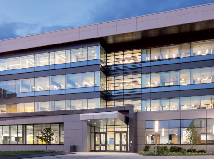 Modern architectural design featuring a multi-level office building with illuminated windows against twilight sky, showcasing reflective glass facades and geometric symmetry with an emphasis on horizontal lines.