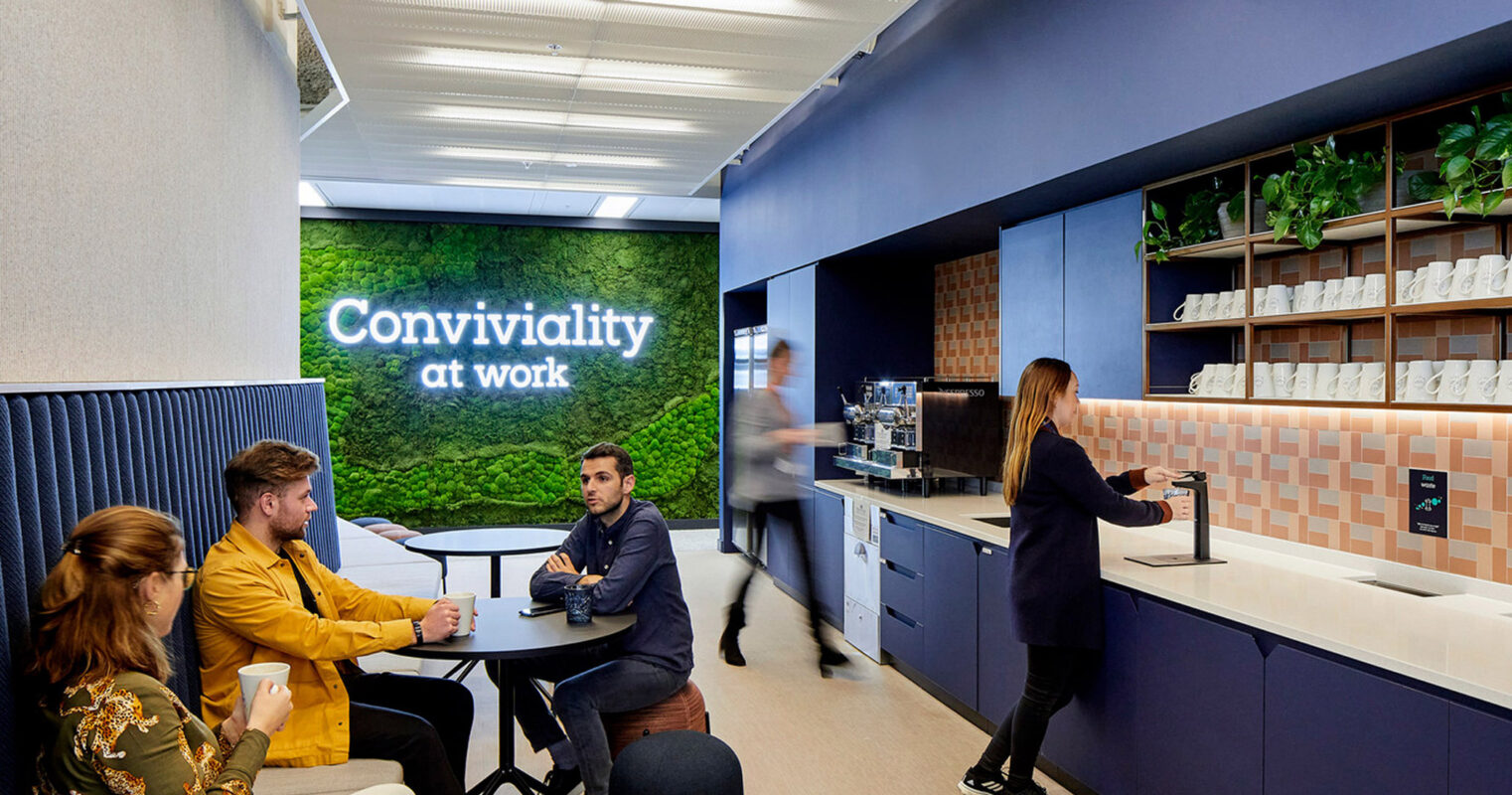 Modern office break room featuring a blue color scheme with integrated plant shelving and a prominent green living wall displaying the text "Conviviality at work". Employees engage casually around tables and the functional kitchenette area.