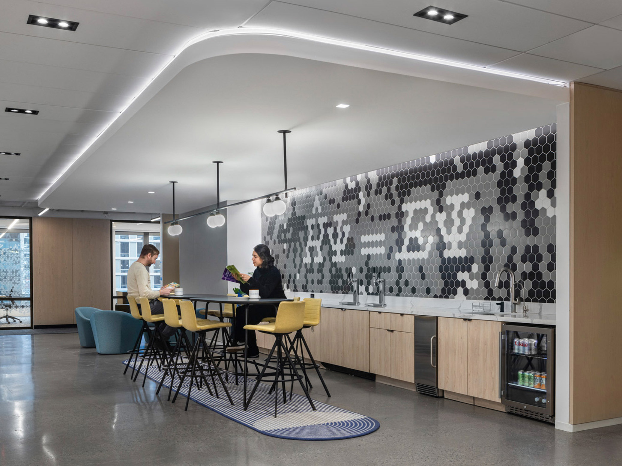 Modern office break room featuring geometric hexagonal tile backsplash, integrated LED lighting in the ceiling, and contemporary furniture with a color palette of blues, yellows, and neutral wood tones.