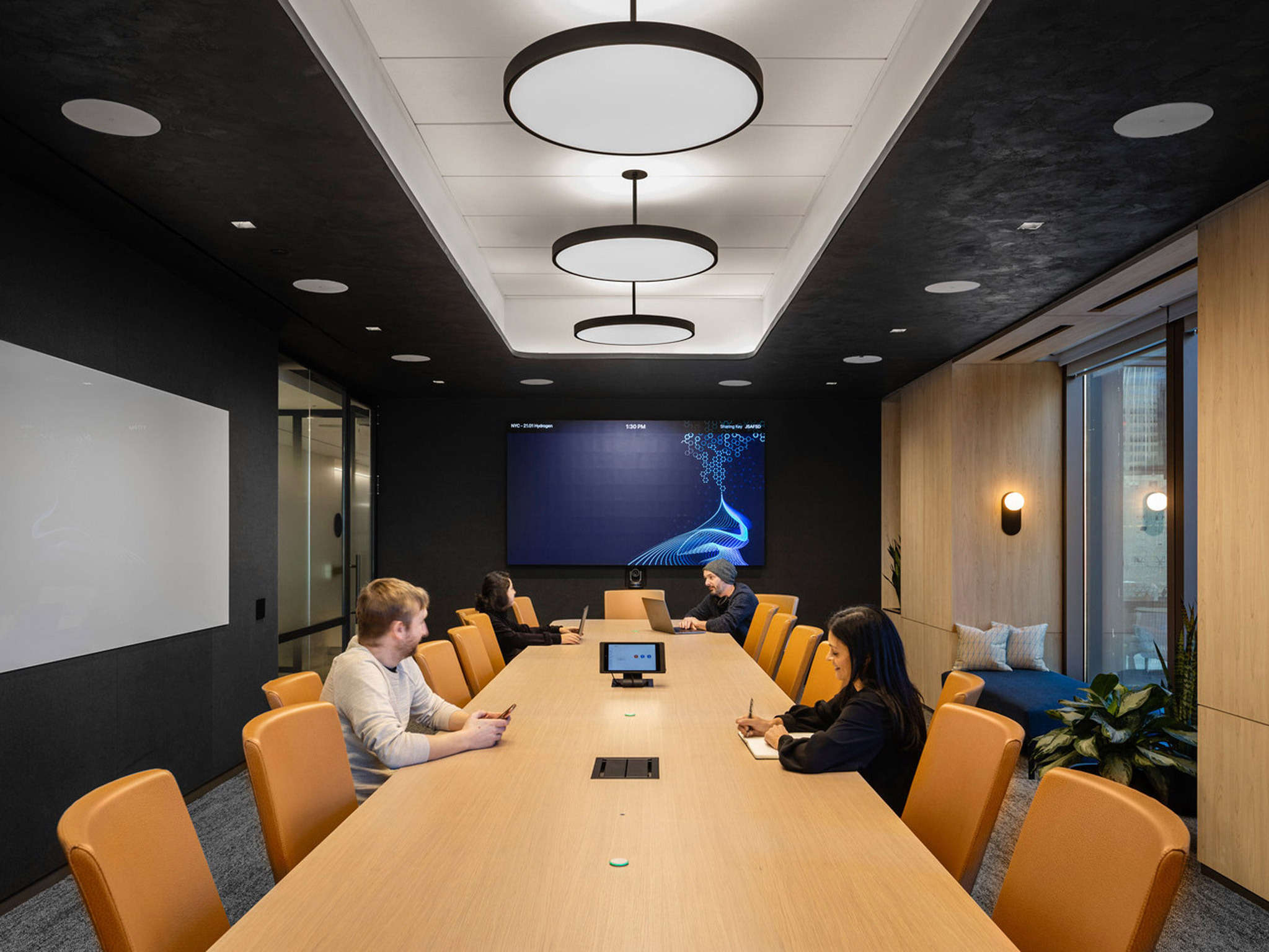 Contemporary boardroom with a sleek, dark ceiling punctuated by circular light fixtures; warm wood paneling intersects with black walls, surrounding a long, tangerine meeting table with attendees in a focused discussion.