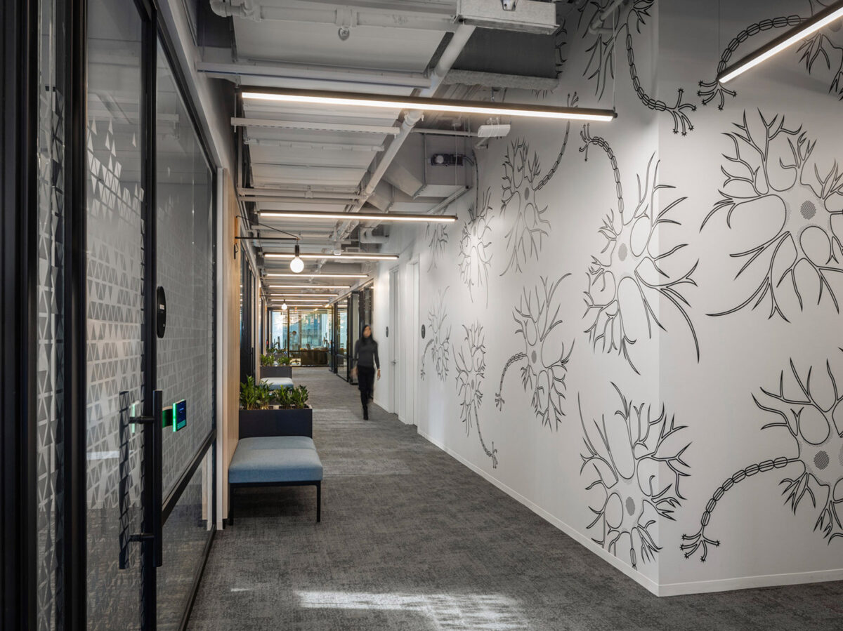 Modern office corridor featuring exposed ceiling utilities and recessed lighting. One wall adorned with a bold floral pattern contrasts with the glass partitions on the opposite side. A blue upholstered bench offers a seating option beside potted greenery, enhancing the workspace's biophilic design.