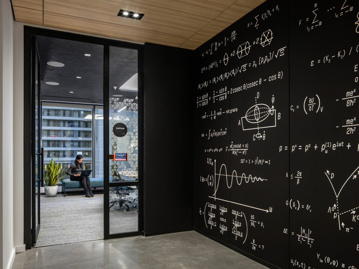 Modern office space featuring a glass wall with a door, leading to an interior adorned with a blackboard wall filled with handwritten mathematical equations. The space balances academic rigor with a casual seating area, reflecting a fusion of collaboration and intellectual pursuit.