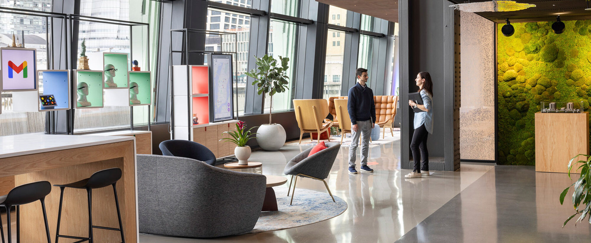 Spacious modern office lounge with floor-to-ceiling windows providing ample natural light. Mixture of casual and formal seating areas with contemporary furniture. Accented by a living green wall, eclectic light fixtures, and colorful art, fostering a creative and collaborative atmosphere. Two individuals converse near a coffee table.