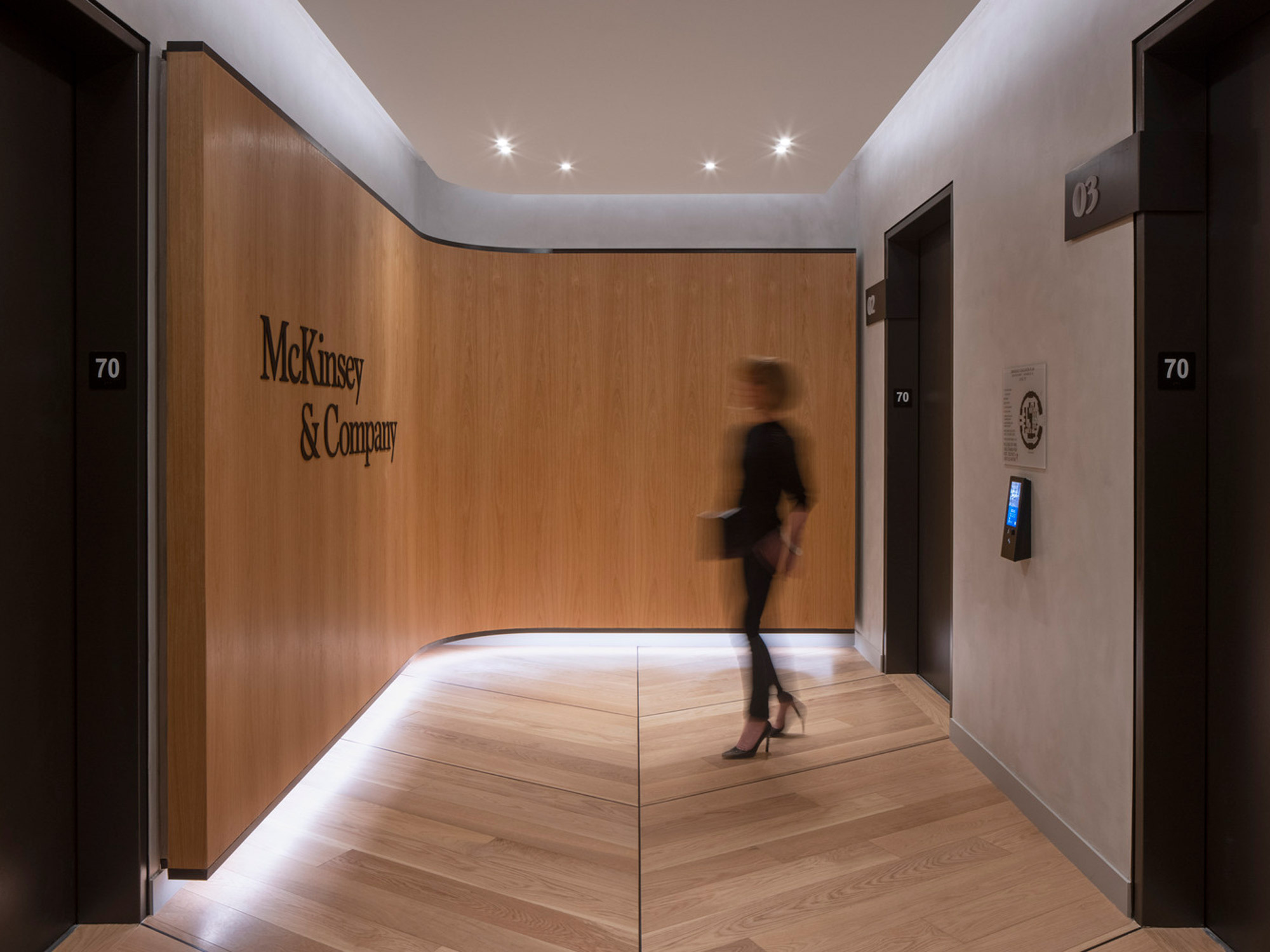 A sleek, modern office corridor featuring warm wooden panels and recessed ceiling lights; signage for 'McKinsey & Company' adorns a door, while a motion blur of a person adds dynamism to the composed setting.