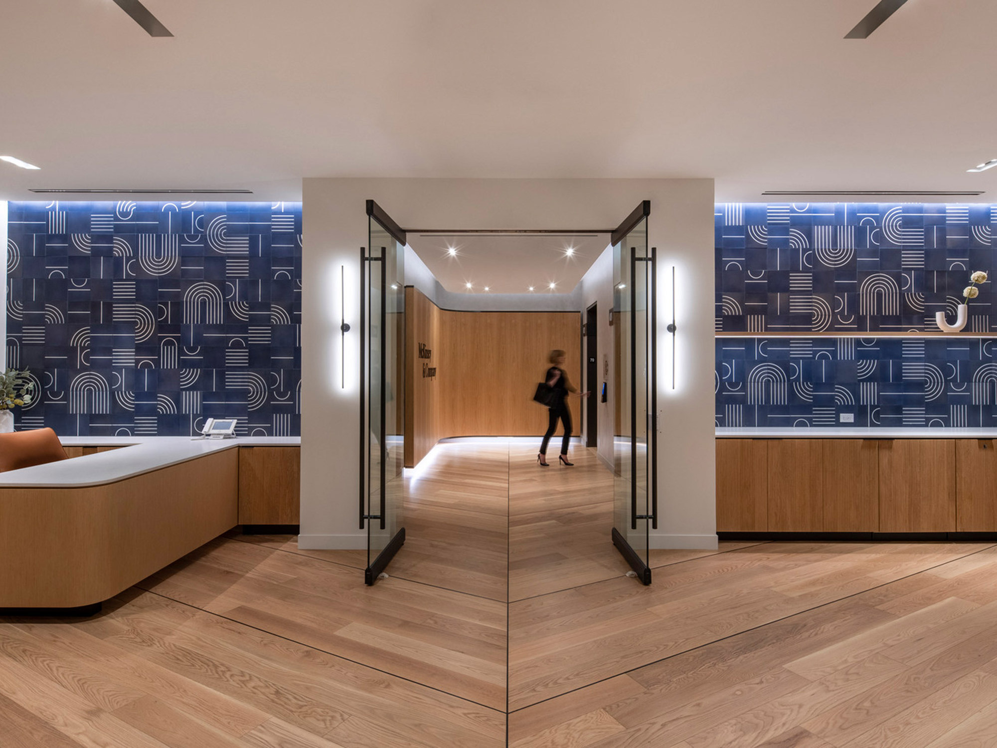 Modern interior showcasing a striking contrast between the bold, geometric-patterned wallpaper and warm-toned, wooden accents along the flooring and curved reception desk. Slim, elegant light fixtures complement the clean lines of the double doors, as a silhouette of a person adds a dynamic presence in the space.