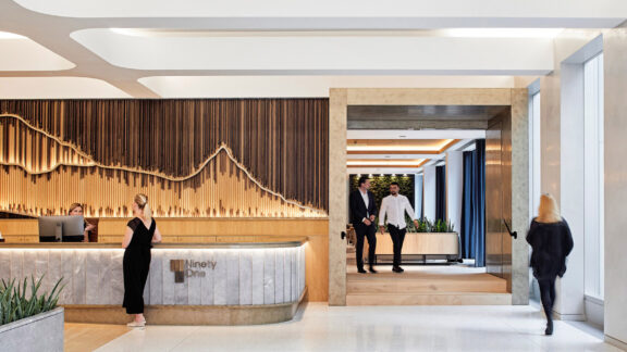 Modern office lobby with a curved wooden reception desk. Marble flooring transitions to a wooden passageway framed by elegant vertical wooden slats. Ambient lighting accentuates the smooth, natural materials and open, welcoming space.