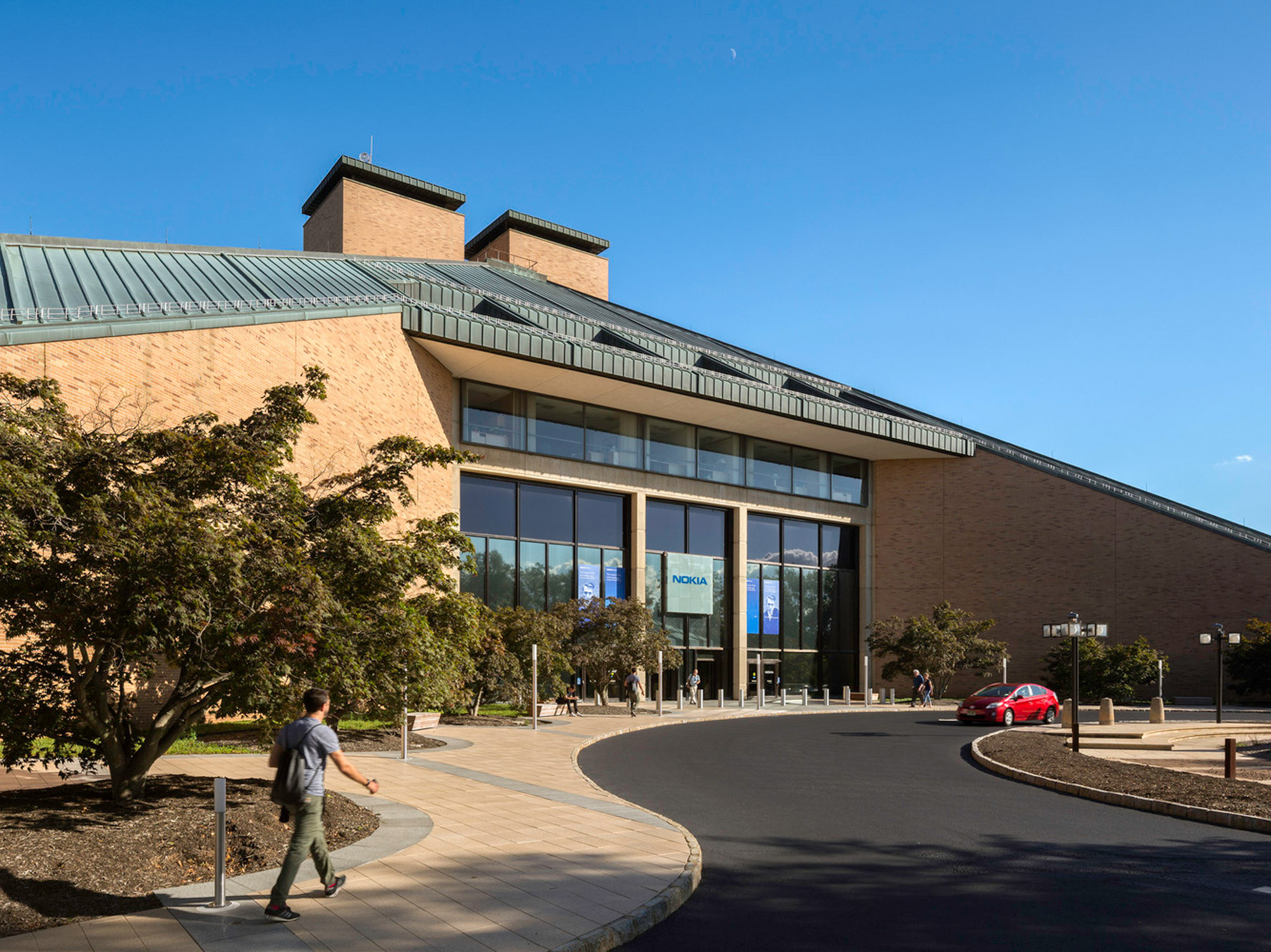 Modern building featuring expansive glass facade and asymmetrical rooflines under clear skies, with a pedestrian pathway leading towards the entrance, surrounded by minimalistic landscaping.