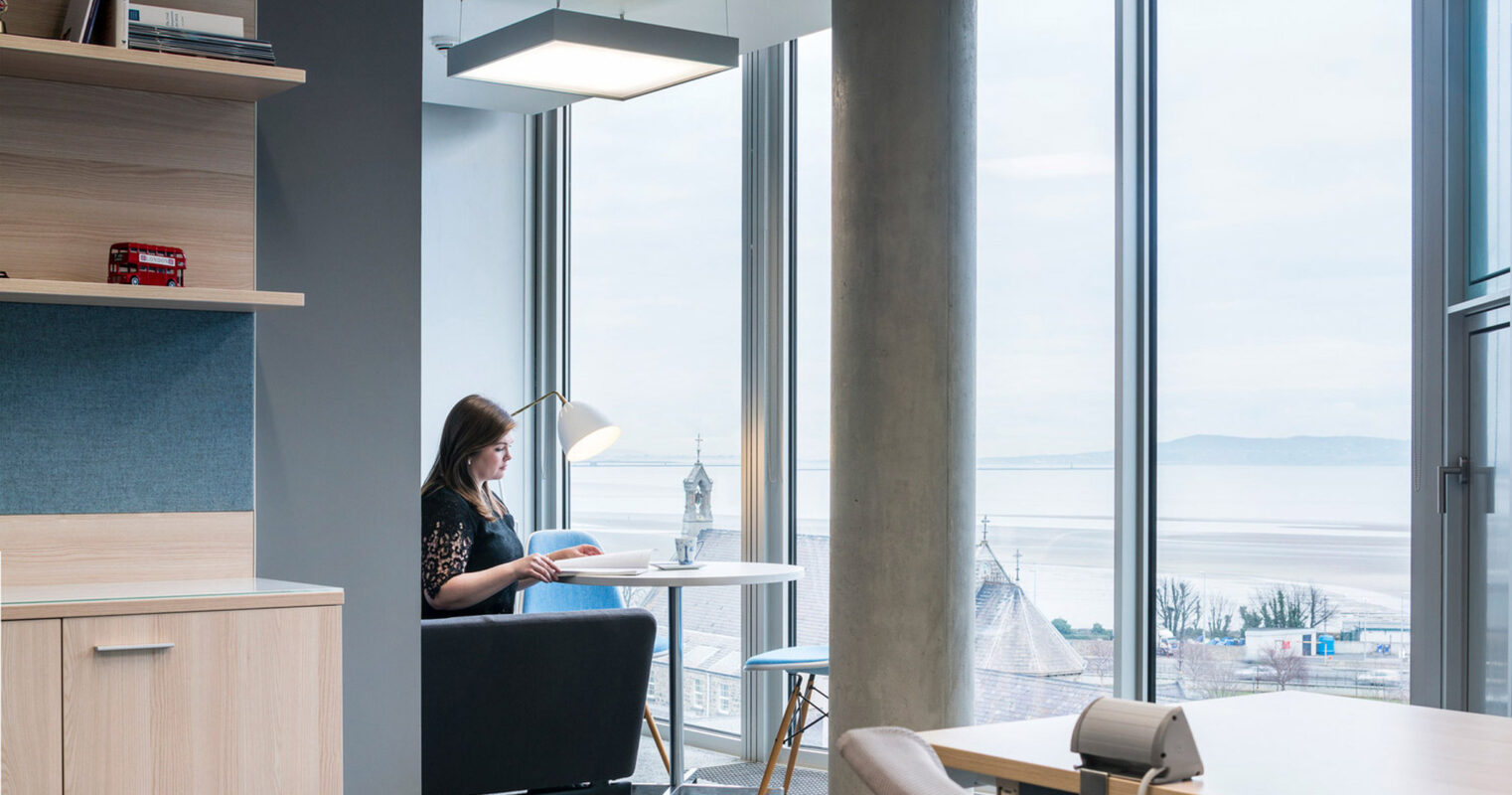 Modern home office with minimalist design, featuring a light wood desk, black chair, and built-in wall shelving. A woman sits reading near a floor-to-ceiling window overlooking a calm water view, illuminated by natural light and a sleek hanging pendant lamp.