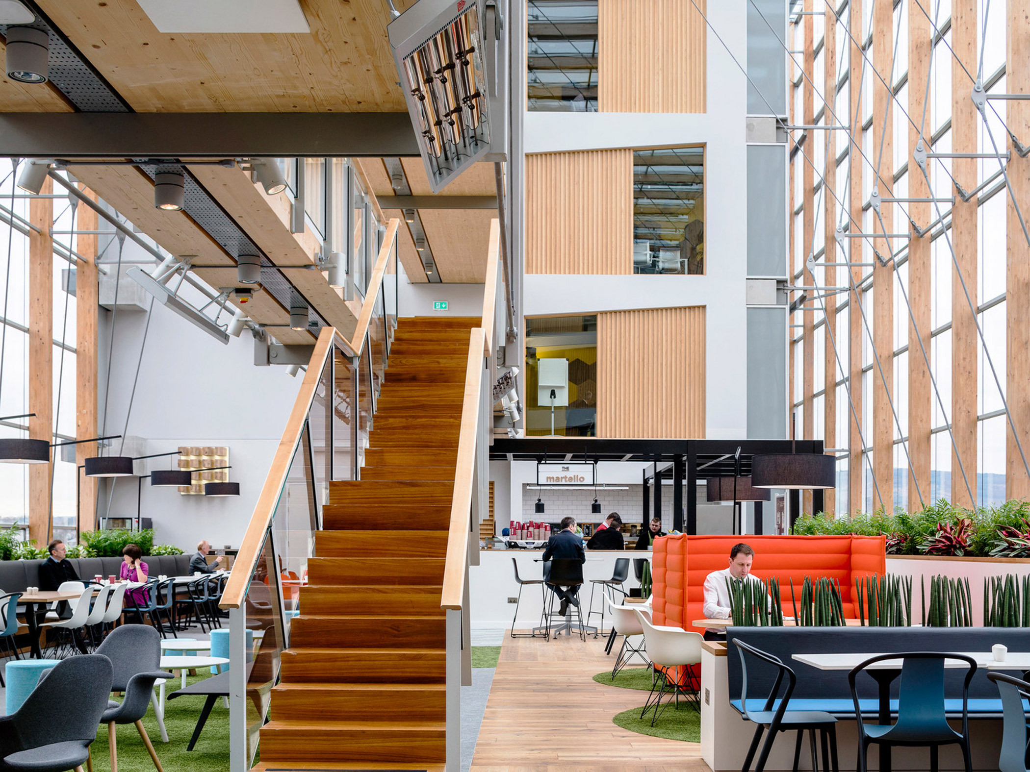 Spacious modern atrium with high ceilings, wooden accents on wall panels and staircases, floor-to-ceiling windows allowing natural light, complemented by contemporary furniture and indoor greenery.