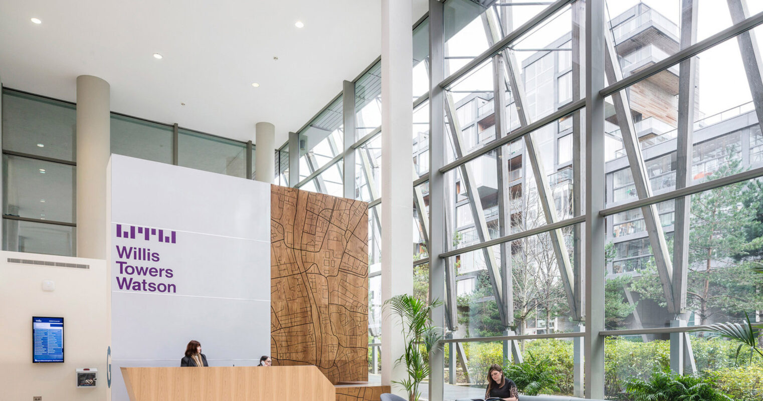 Spacious atrium lobby featuring high ceilings with geometric skylights, a large scale wood-paneled reception desk, sleek gray sofas on patterned area rugs, and airy greenery, embodying modern corporate design.