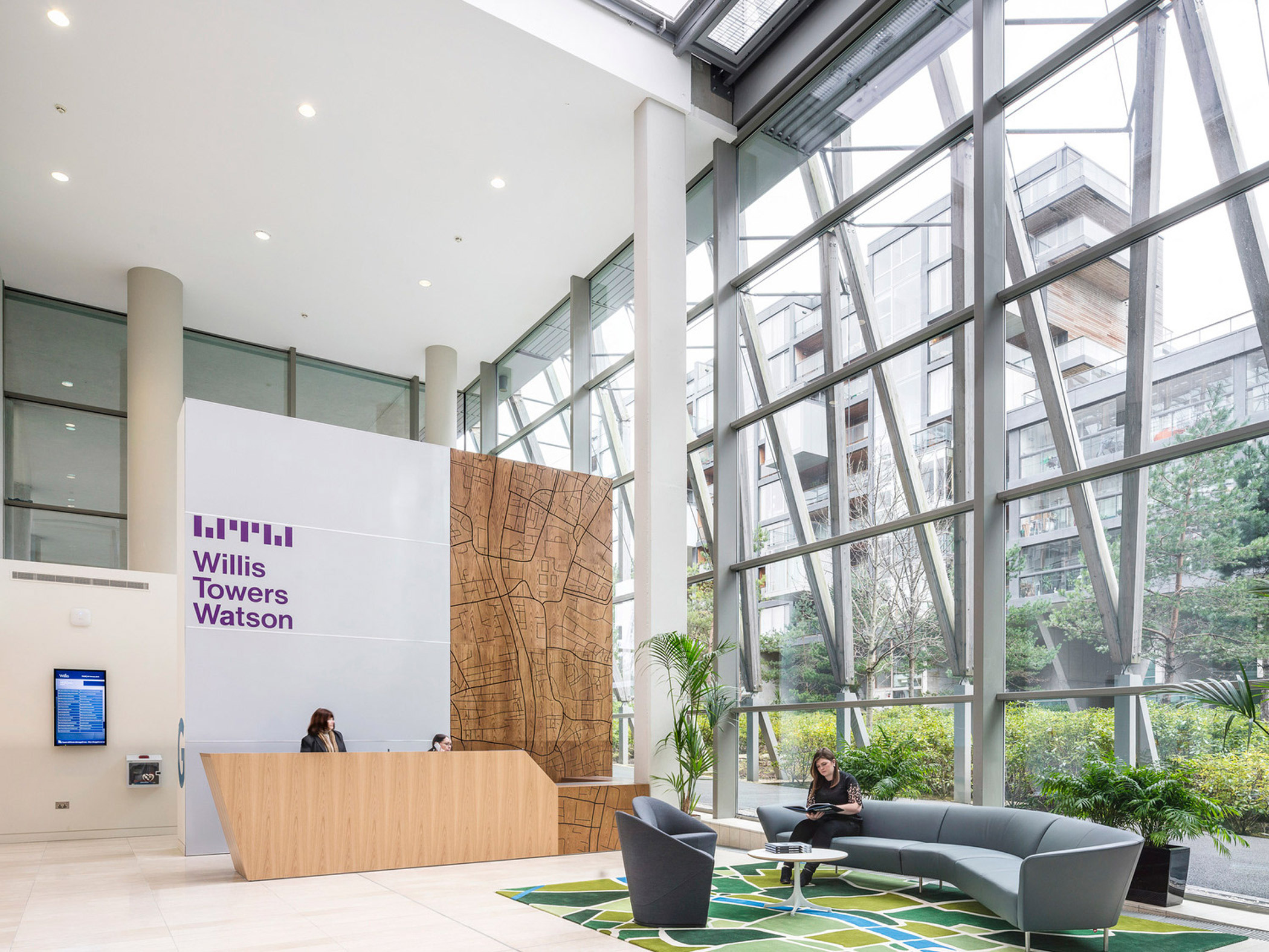 Spacious atrium lobby featuring high ceilings with geometric skylights, a large scale wood-paneled reception desk, sleek gray sofas on patterned area rugs, and airy greenery, embodying modern corporate design.