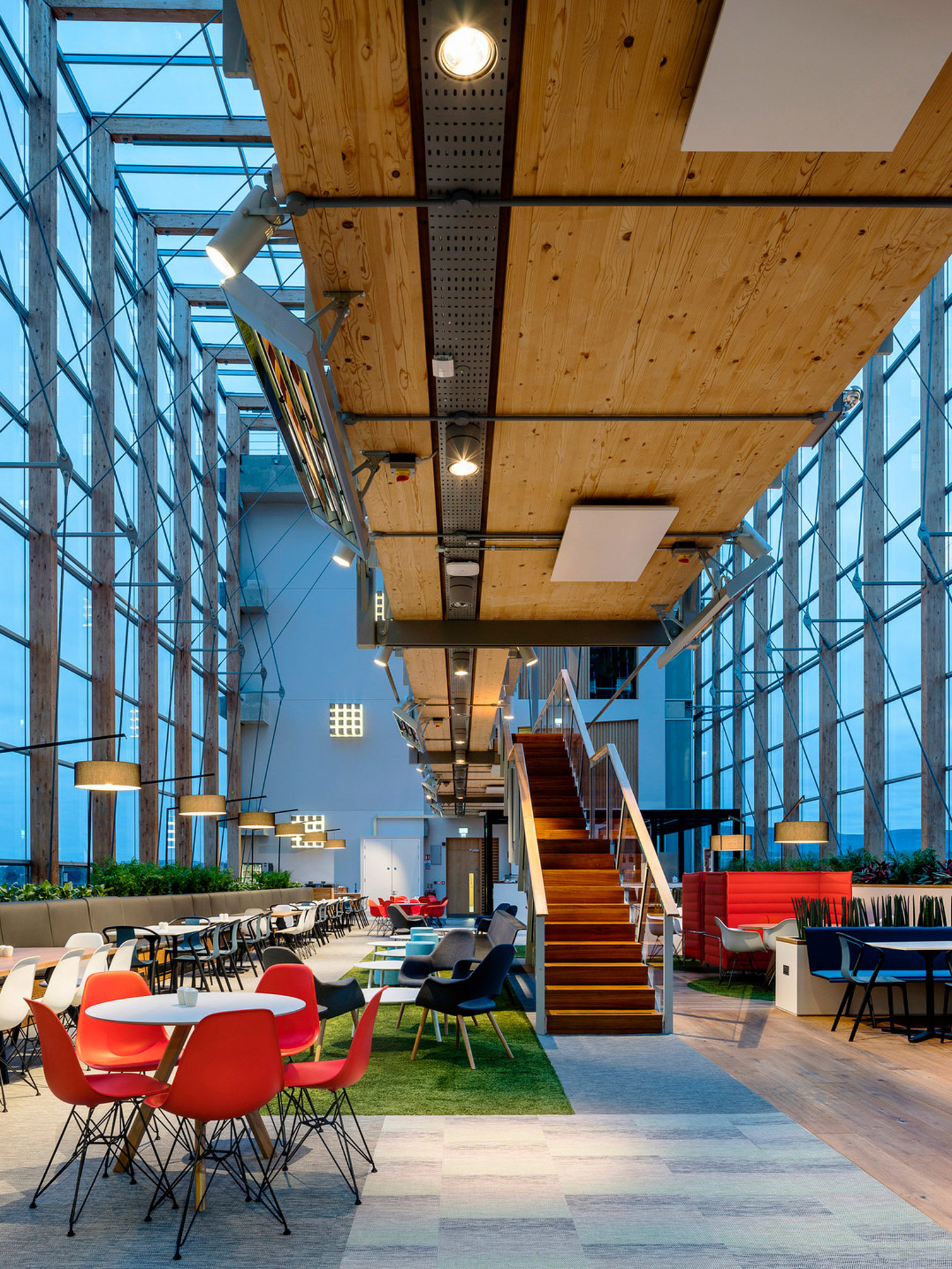 Spacious modern office atrium with high ceilings, wooden beams, and expansive windows. A mix of red and white chairs are paired with natural wood tables. A wooden staircase with glass balustrade leads to the upper level, enhancing the airy, open-plan design.