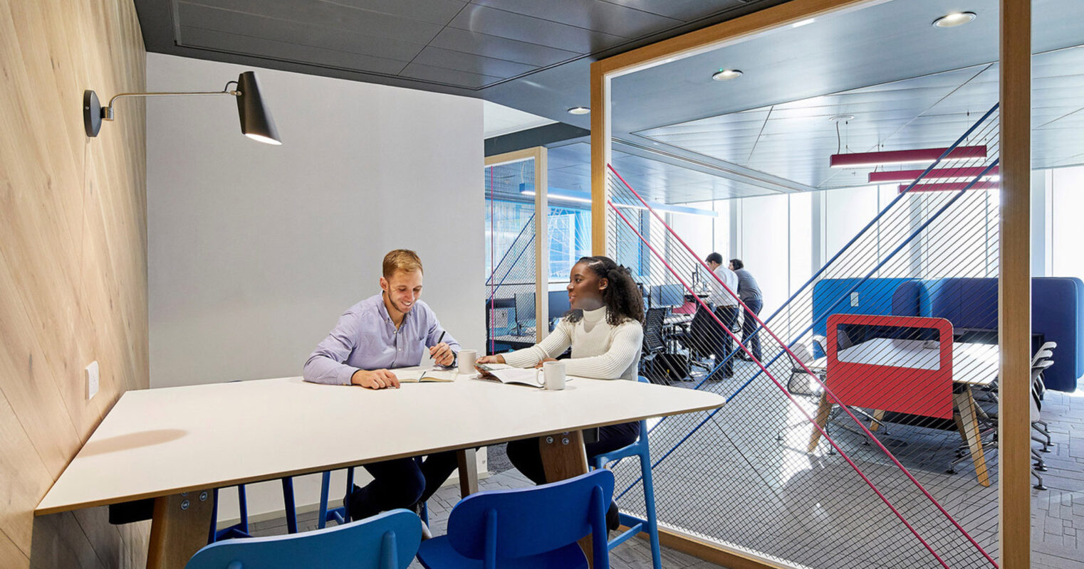 Modern office space featuring a minimalist design with a masculine palette, accentuated by vibrant blue chairs and a bold red geometric room divider that creates a dynamic visual contrast, promoting an energetic and collaborative work environment.