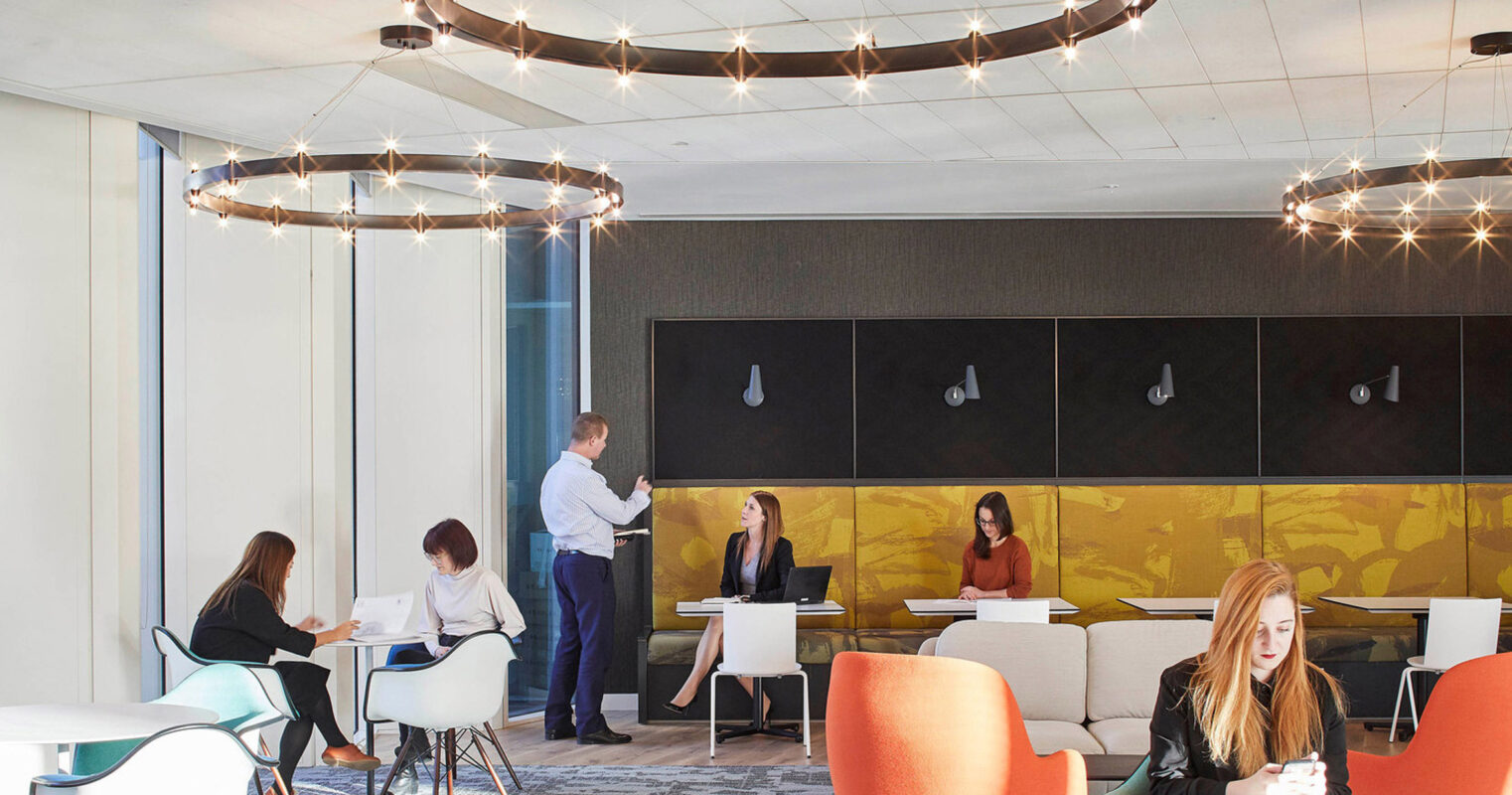 Modern open-plan office space with eclectic seating arrangements from individual tables to casual lounge areas, accented by vibrant orange chairs and understated grey tones. An impressive circular lighting fixture anchors the room, complementing the natural light from floor-to-ceiling windows, while employees engage in various work activities.