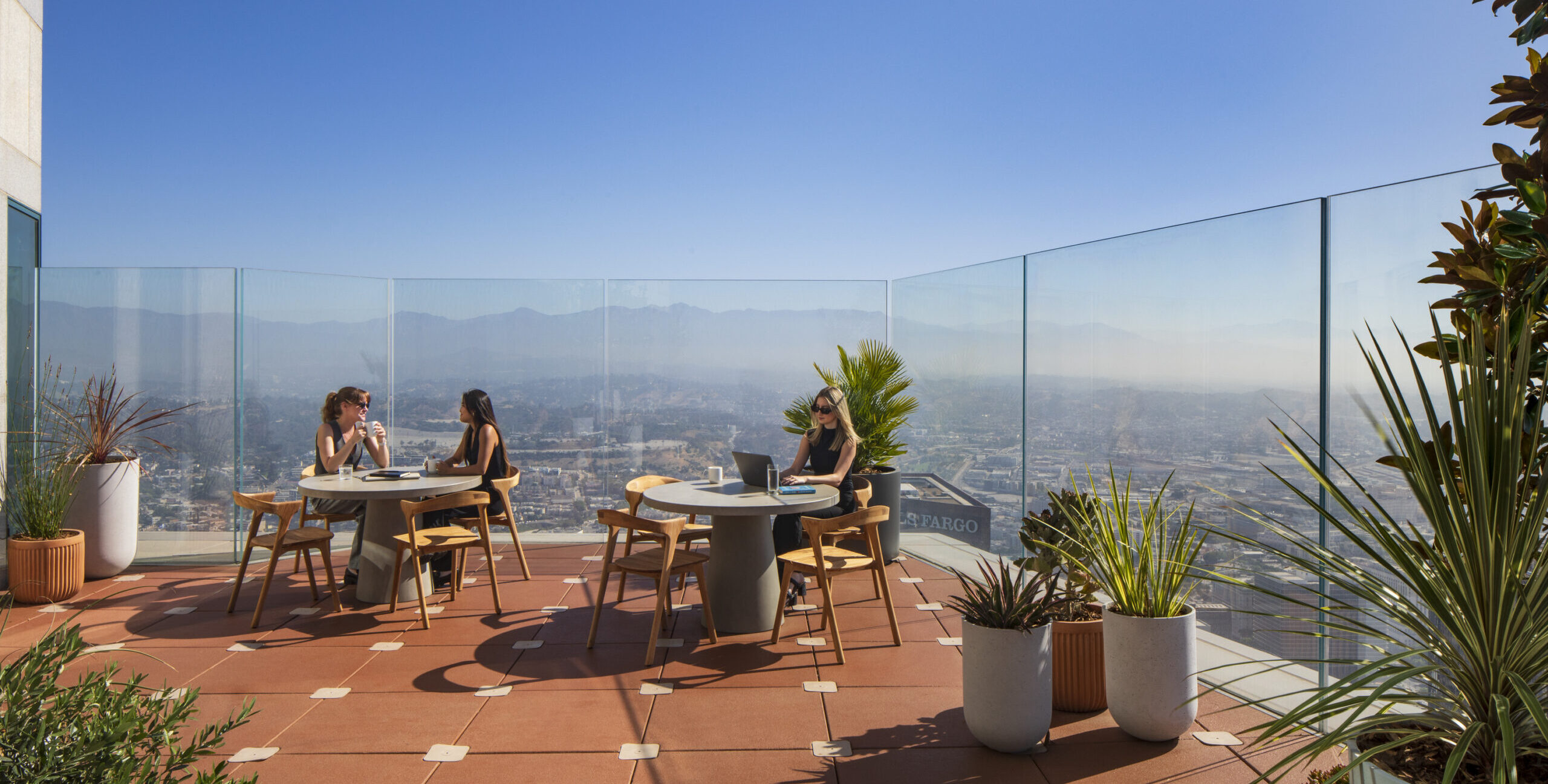 A modern rooftop terrace features sleek wooden furniture and contemporary planters filled with lush greenery. Panoramic glass panels offer breathtaking views of the cityscape and mountains. Two women enjoy their time, highlighting the terrace's inviting atmosphere and the seamless integration of outdoor living with urban life.