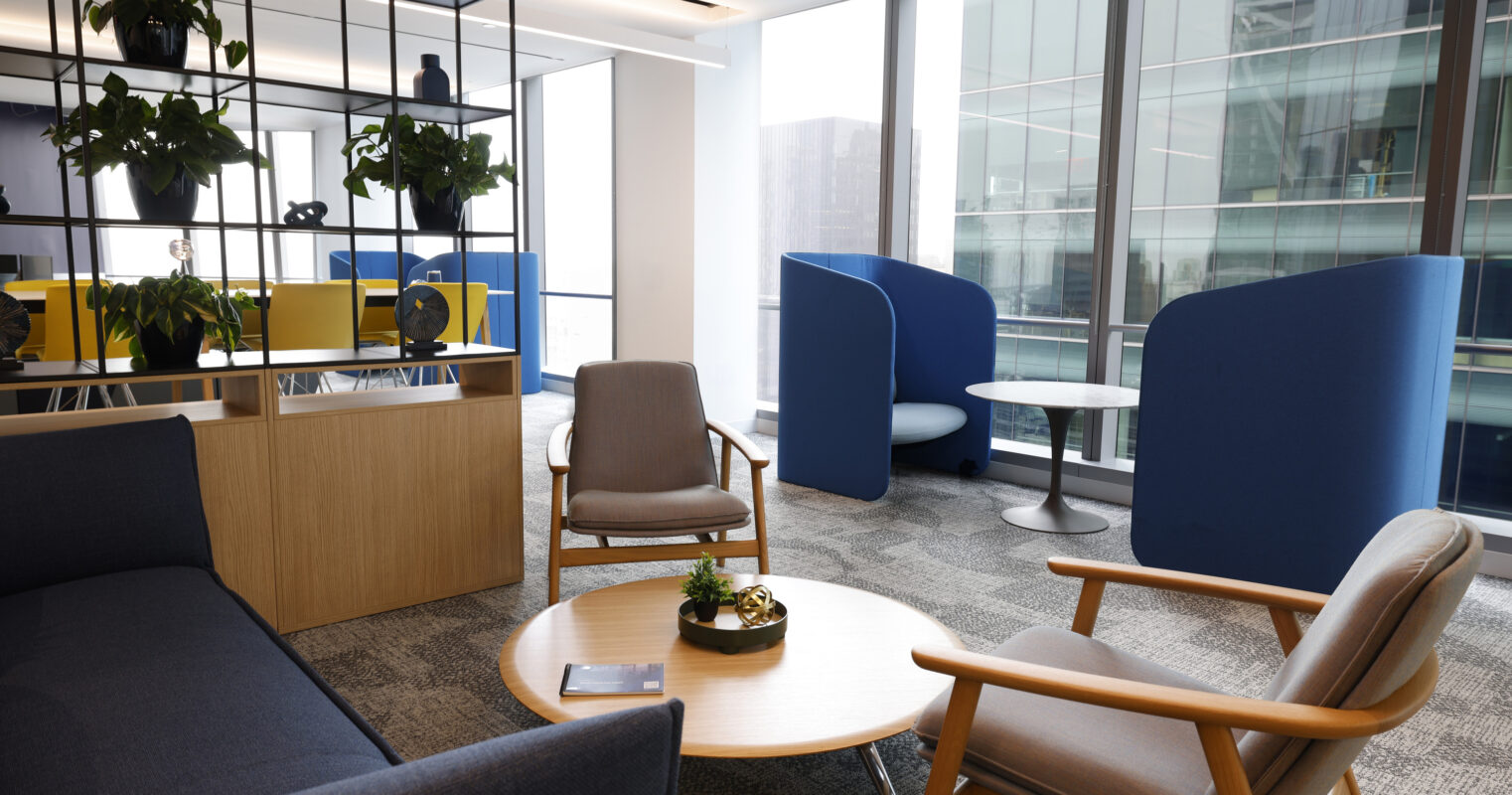 Modern office lounge featuring a mix of soft furnishings and collaborative workspaces. A cozy sofa with ergonomic chairs surrounds a wooden coffee table, complemented by greenery on display. Floor-to-ceiling windows flood the area with natural light, while privacy pods create informal meeting spots, enhancing workspace functionality and aesthetic appeal.