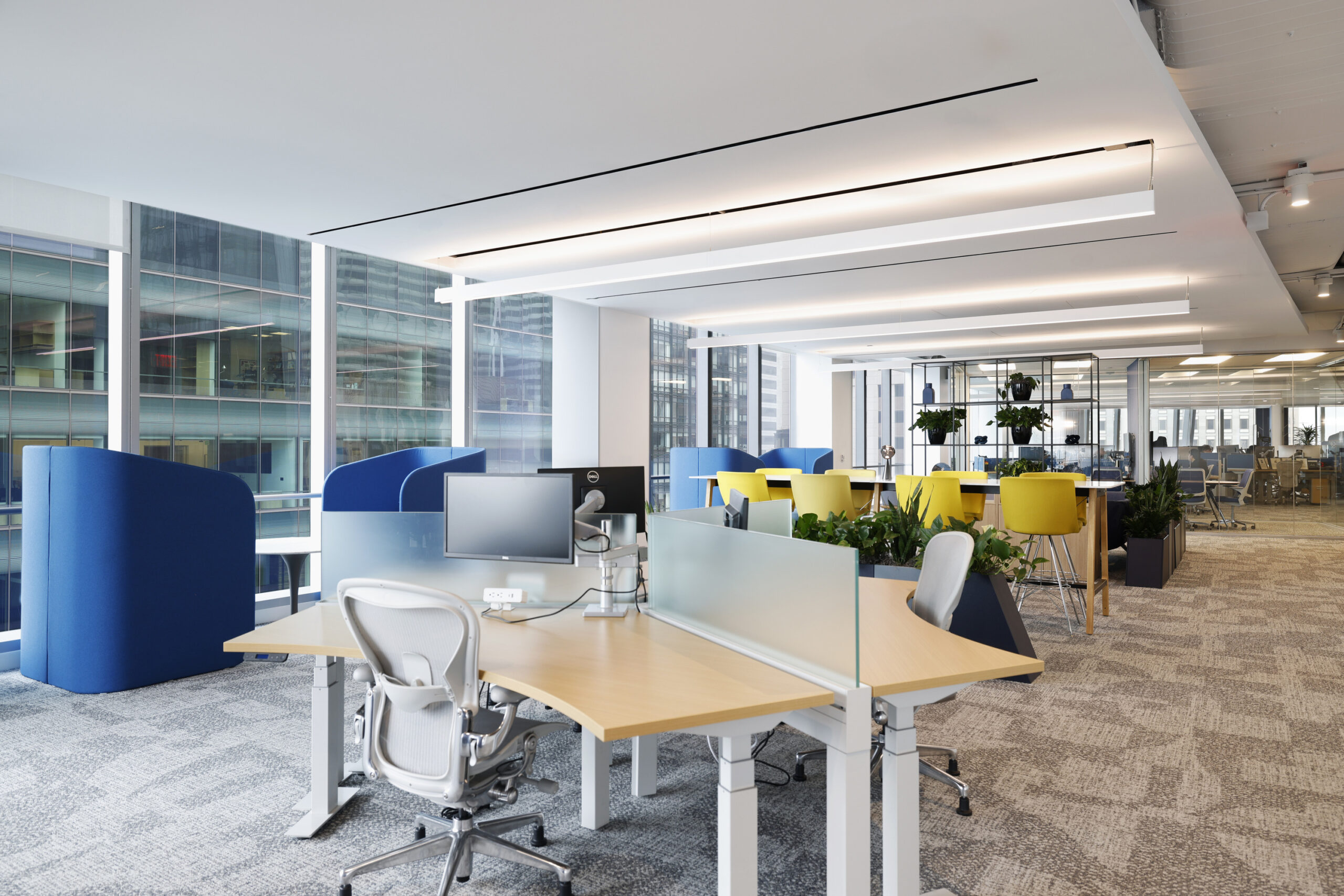 Modern office space designed for collaboration, featuring open workstations with ergonomic chairs and adjustable desks. Bright, natural light floods the area through large windows, complemented by vibrant, colorful seating pods. The incorporation of greenery adds a touch of nature, enhancing creativity and well-being within the workplace.