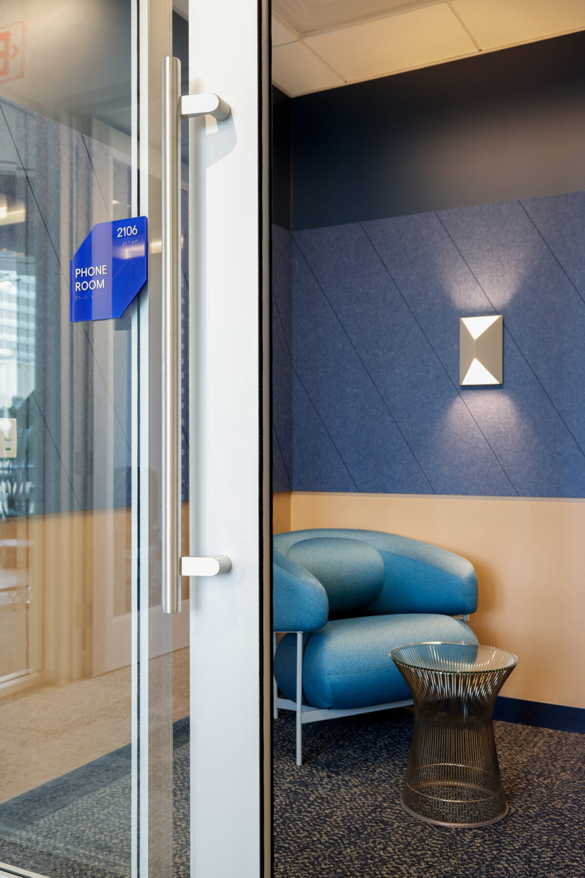 A modern phone room featuring a curved blue accent chair and a sleek metallic side table. The interior walls are decorated with a geometric blue and beige color scheme, complemented by stylish wall lighting. The glass door provides a minimalist entryway, enhancing the space's contemporary feel.