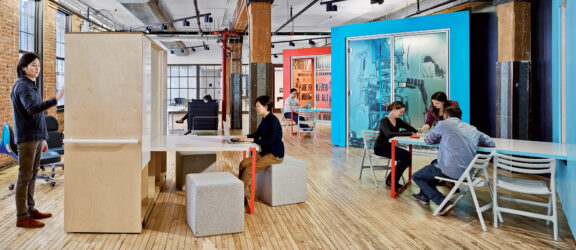 Open-plan office space blending modern and industrial elements, featuring exposed brick and wooden beams, vibrant teal office pods with frosted glass, hardwood floors, and a mix of seated and standing workspace options. Employees actively engage in collaborative and individual tasks.